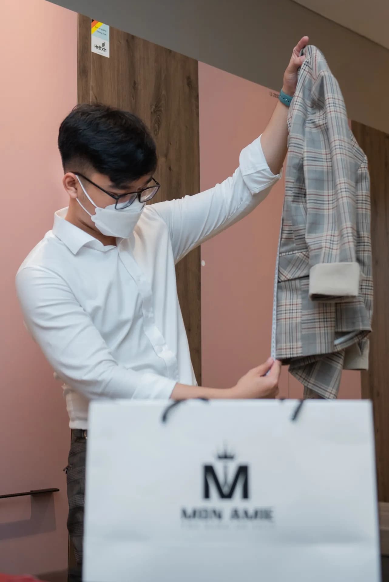 Nhân viên giơ lên chiếc áo blazer kẻ caro xám bên túi Mon Amie, nền hồng nhẹ