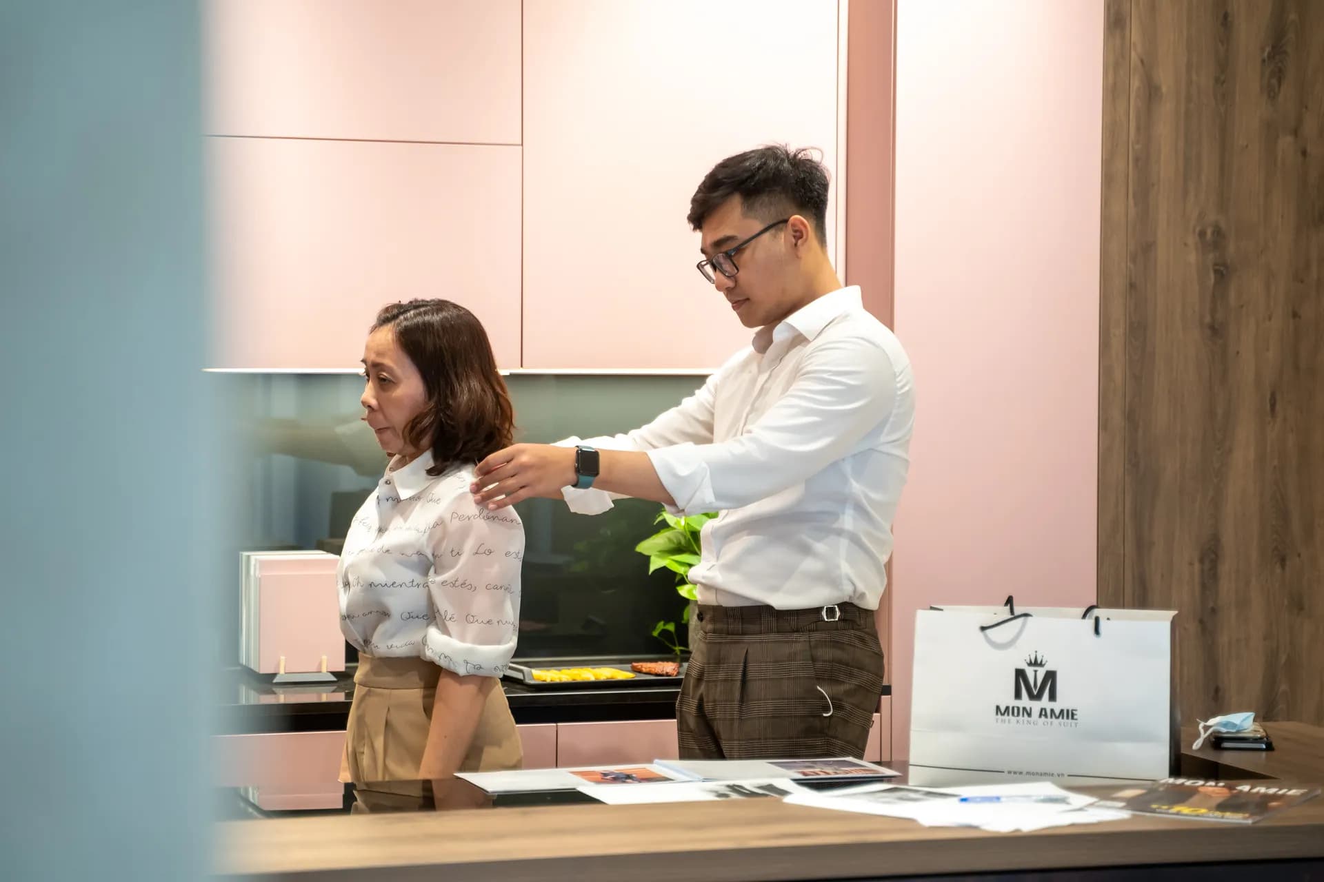 Đo vai cho khách nữ giữa gian bếp tông hồng, túi Mon Amie trên bàn