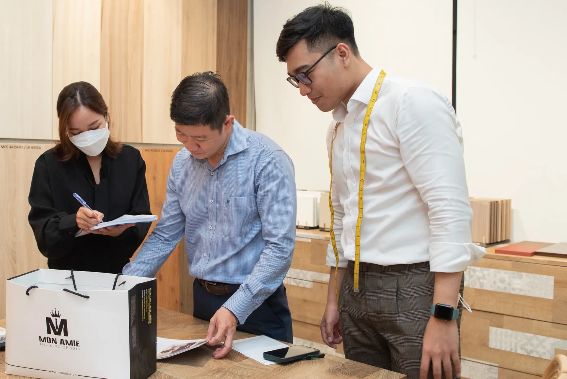 Thợ đo thước dây vàng vắt cổ cùng khách và trợ lý ghi số đo bên túi Mon Amie