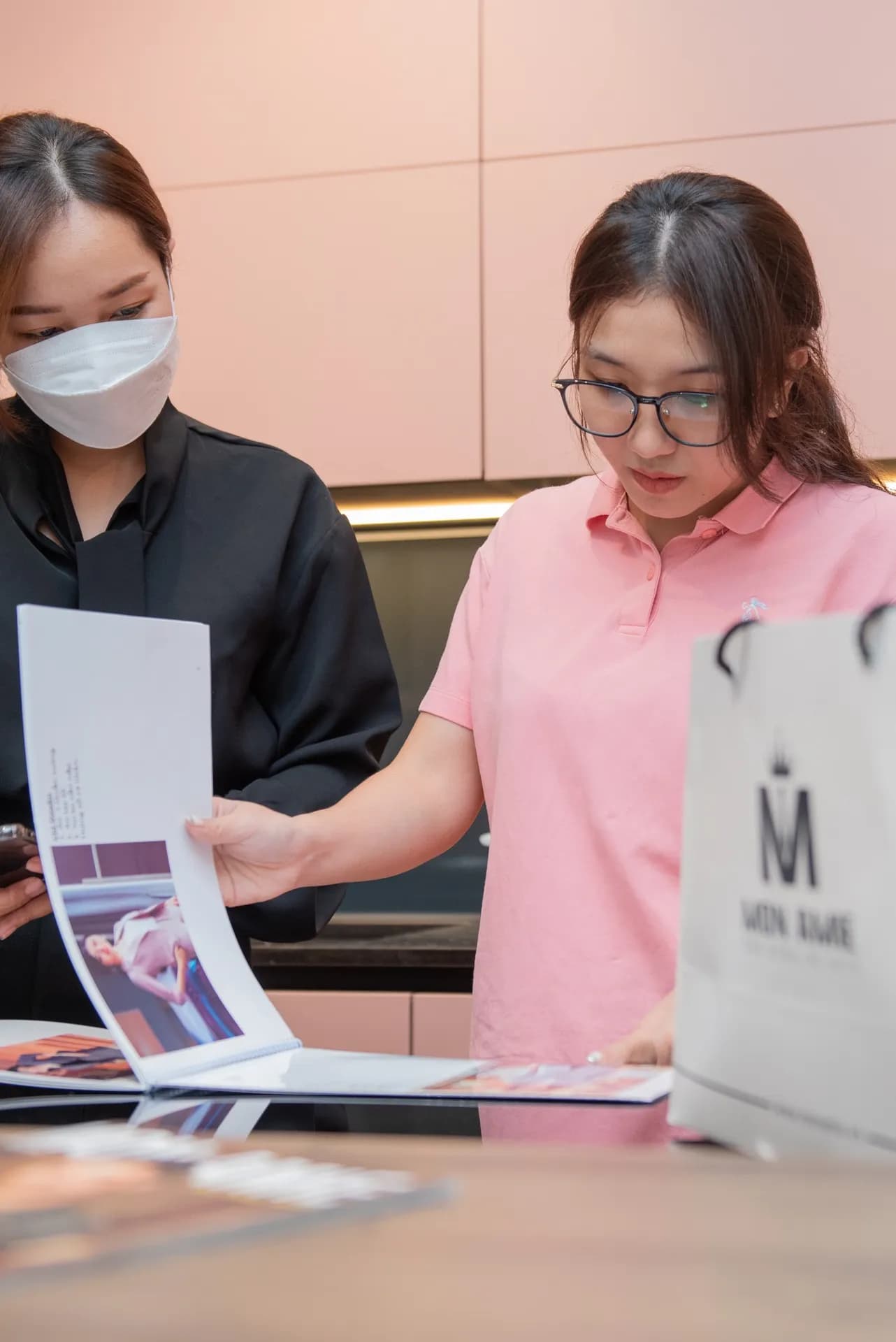 Khách nữ áo polo hồng lật xem lookbook Mon Amie cùng nhân viên tư vấn