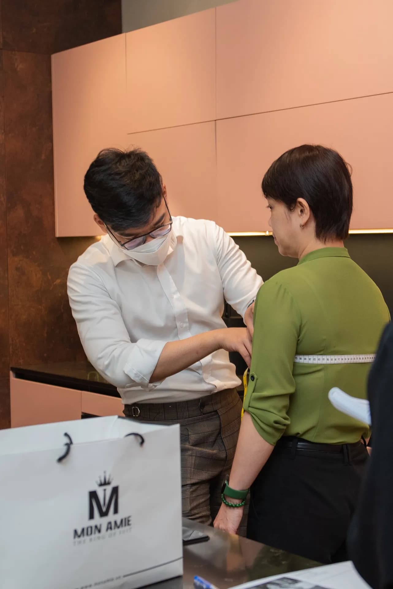 Đo vòng ngực cho khách nữ áo xanh rêu, túi Mon Amie ở tiền cảnh, nền tủ hồng