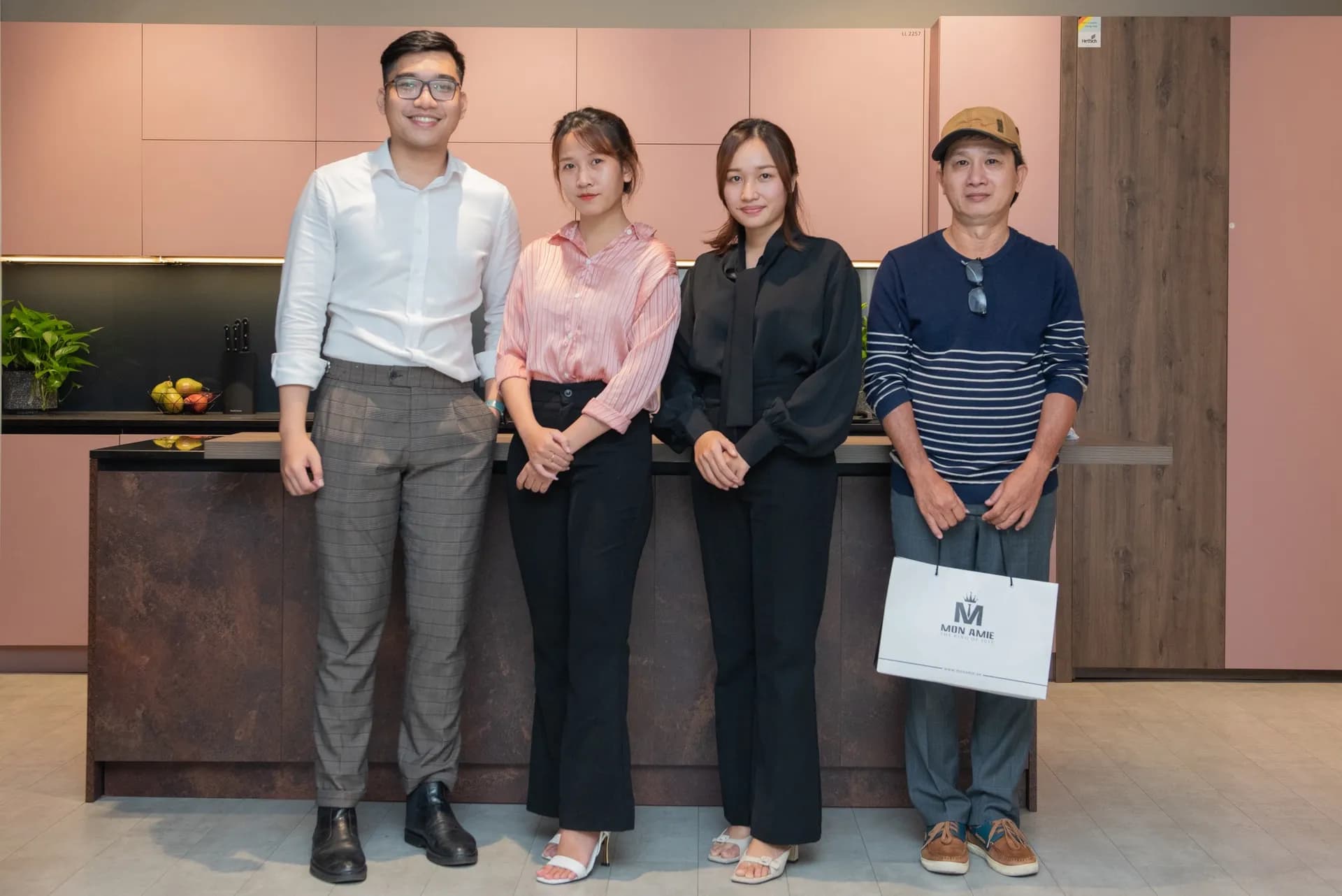 Ảnh lưu niệm bốn người sau buổi đo, một khách cầm túi Mon Amie trước tủ bếp hồng
