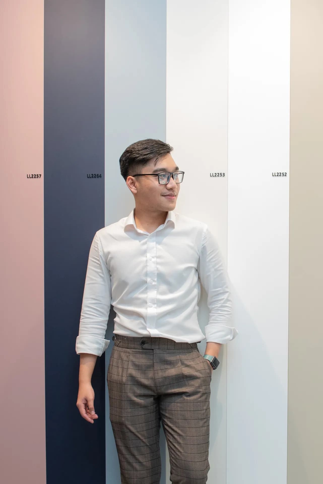 Sơ mi trắng quần tây caro nâu, quay nghiêng cười nhẹ trước vách màu pastel