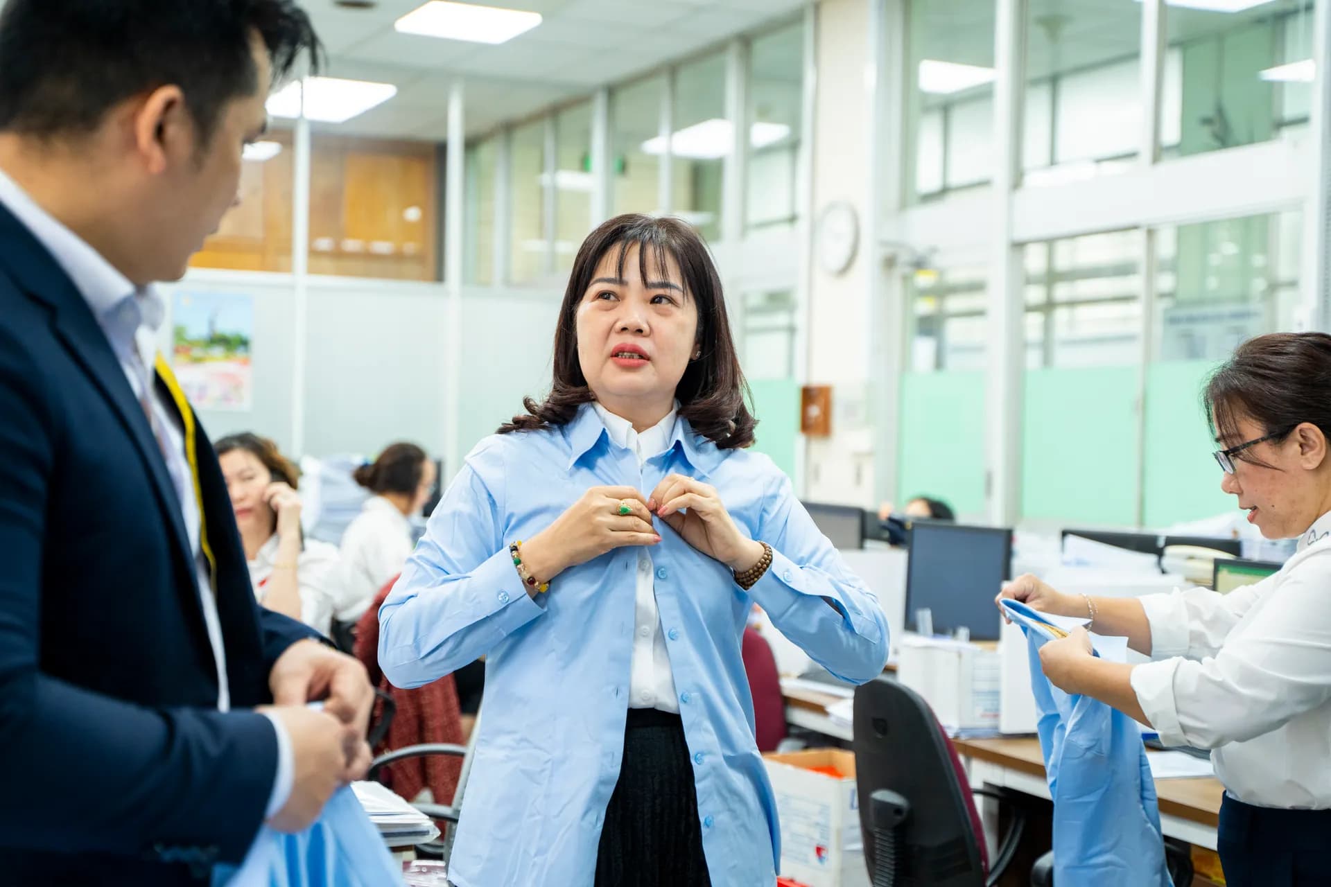 Nhân viên nữ mỉm cười cài khuy áo sơ mi xanh nhạt vừa nhận, thợ may đứng cạnh