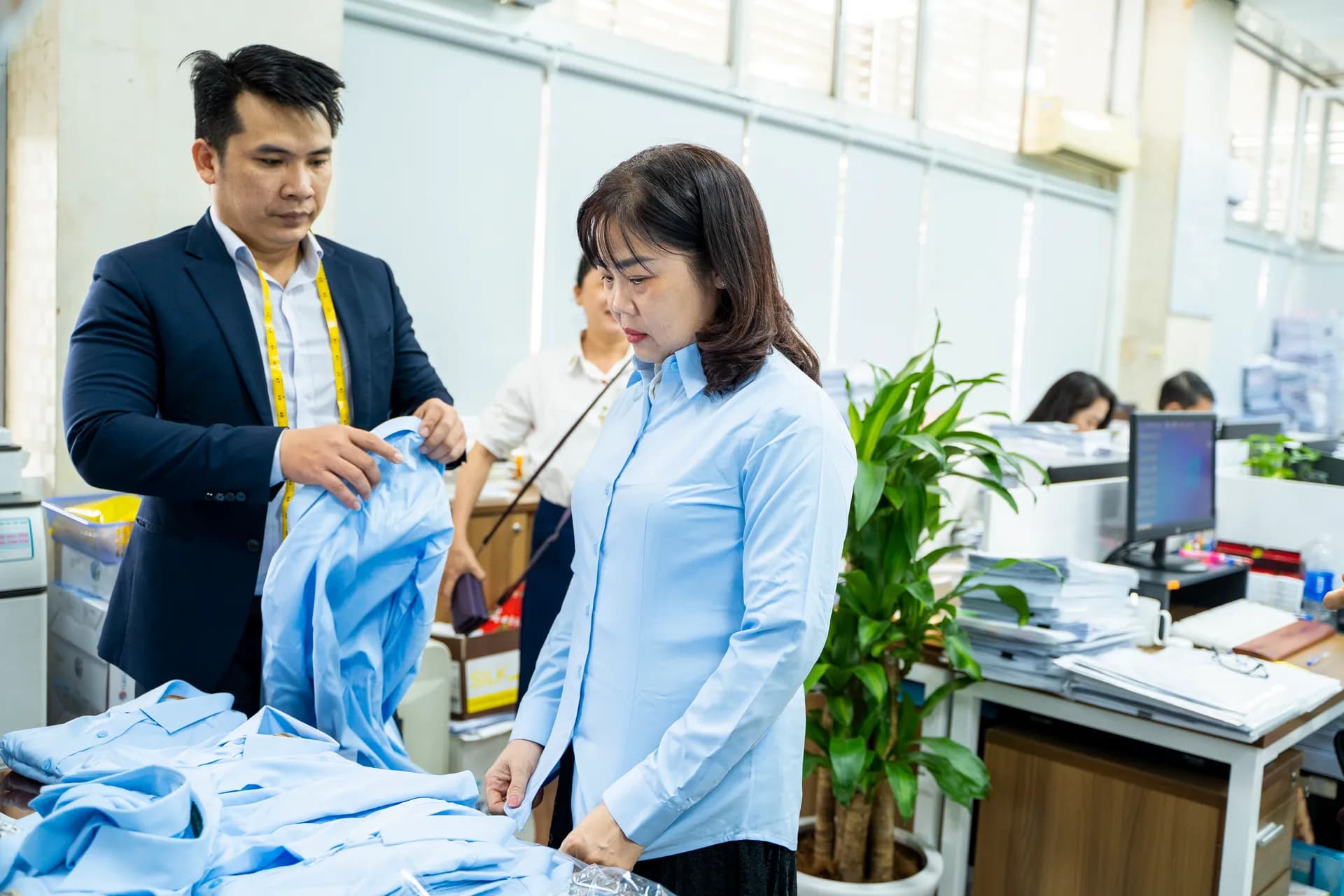 Thợ may vest navy cầm sơ mi xanh nhạt, khách nữ mặc thử trong văn phòng sáng