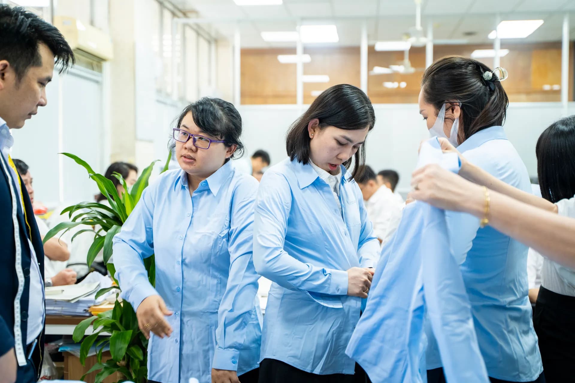 Các nhân viên nữ mặc thử sơ mi đồng phục xanh nhạt, chỉnh khuy trước đồng nghiệp