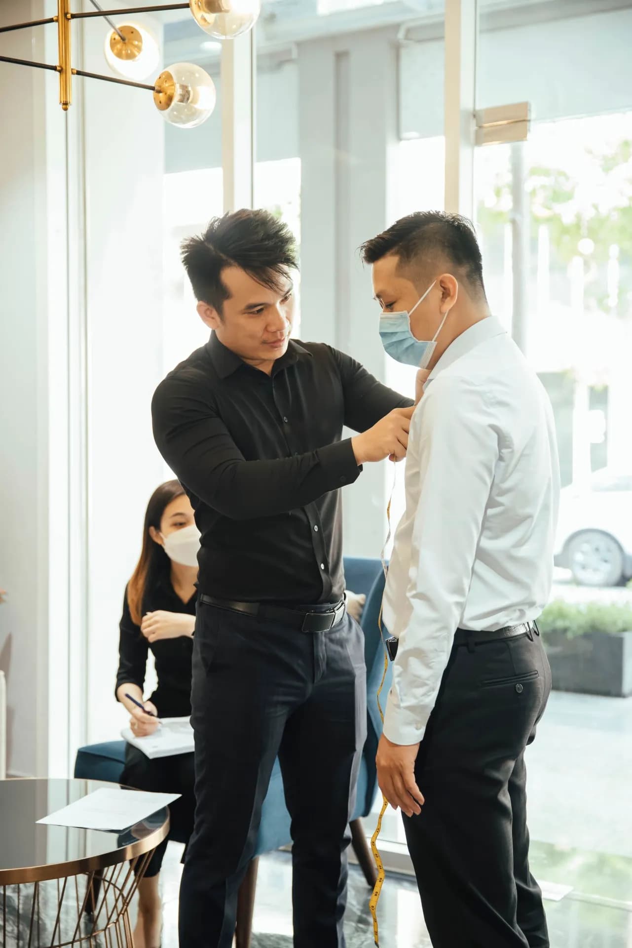 Thợ may áo đen đo vai cho khách sơ mi trắng bên cửa kính ngập nắng