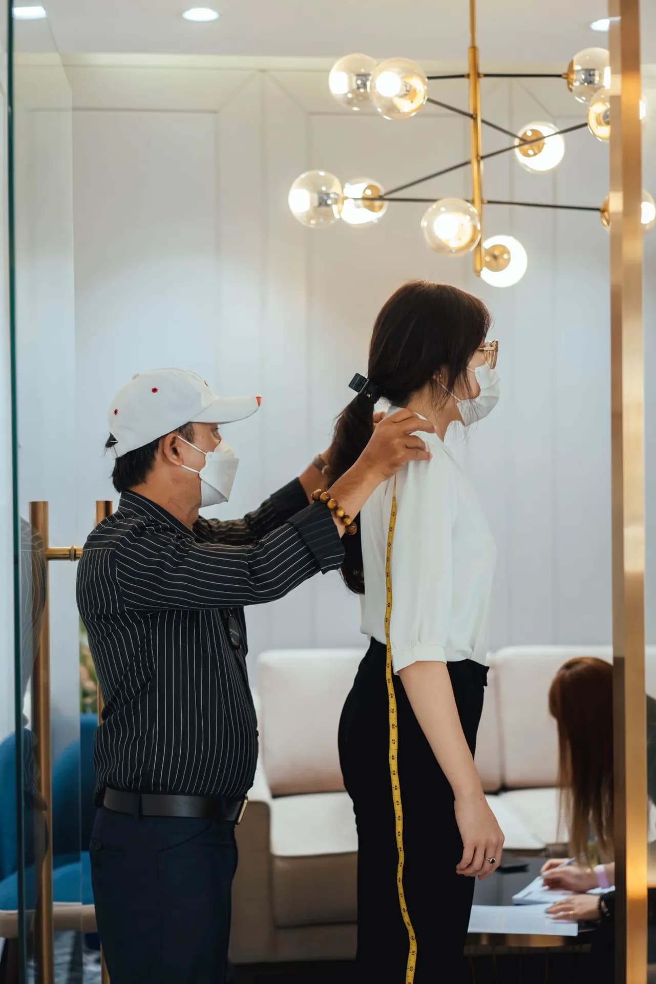 Thợ may áo sọc đen đo chiều dài lưng khách nữ, đèn chùm vàng phía sau