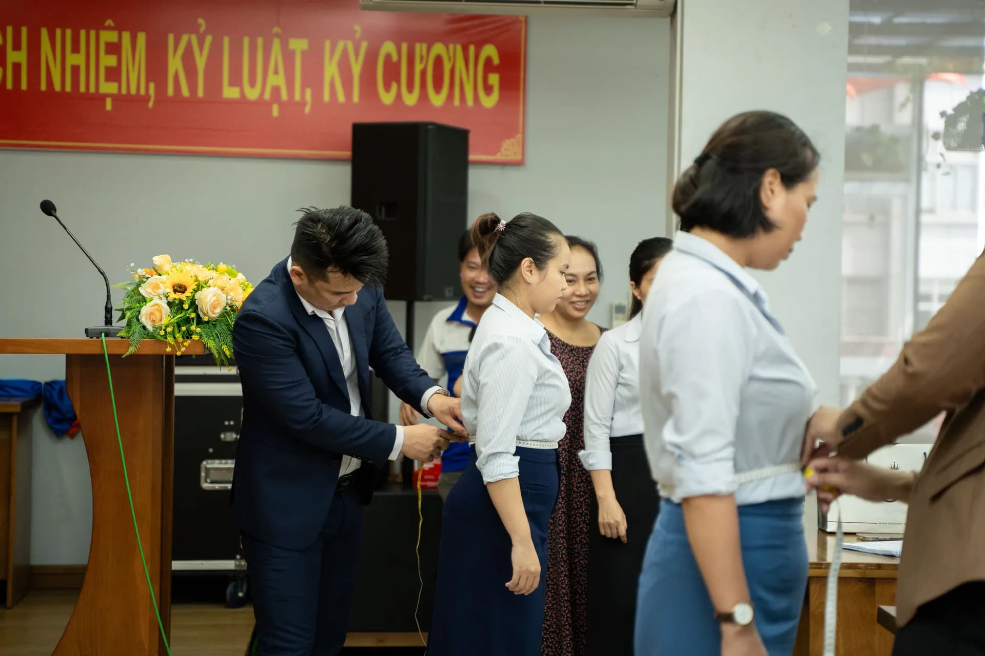 Đo vòng eo cho nhân viên nữ bên bục hội trường, hàng người chờ phía sau
