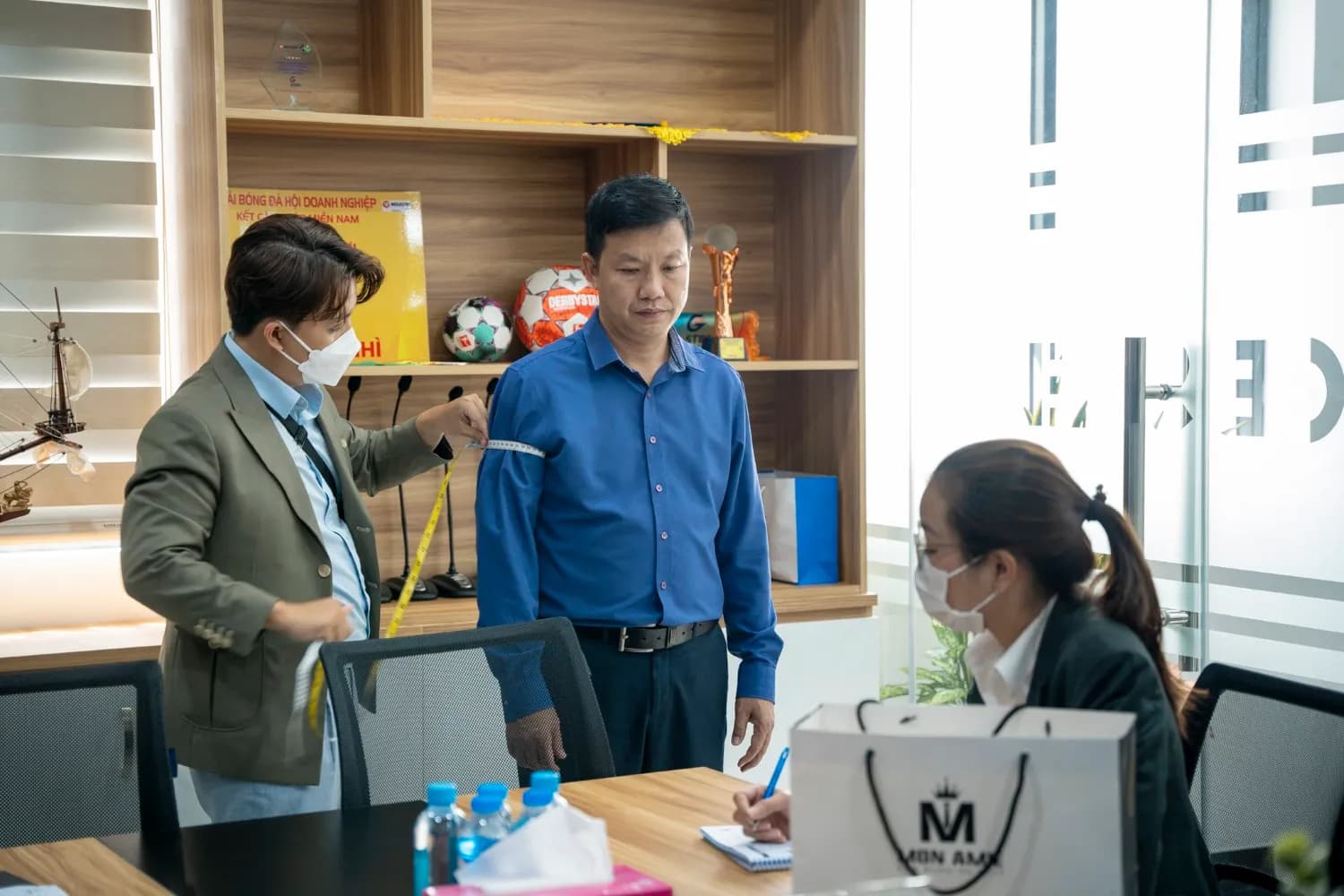 Đo tay áo cho khách sơ mi xanh trong phòng họp, thư ký ghi số đo, túi Mon Amie trên bàn