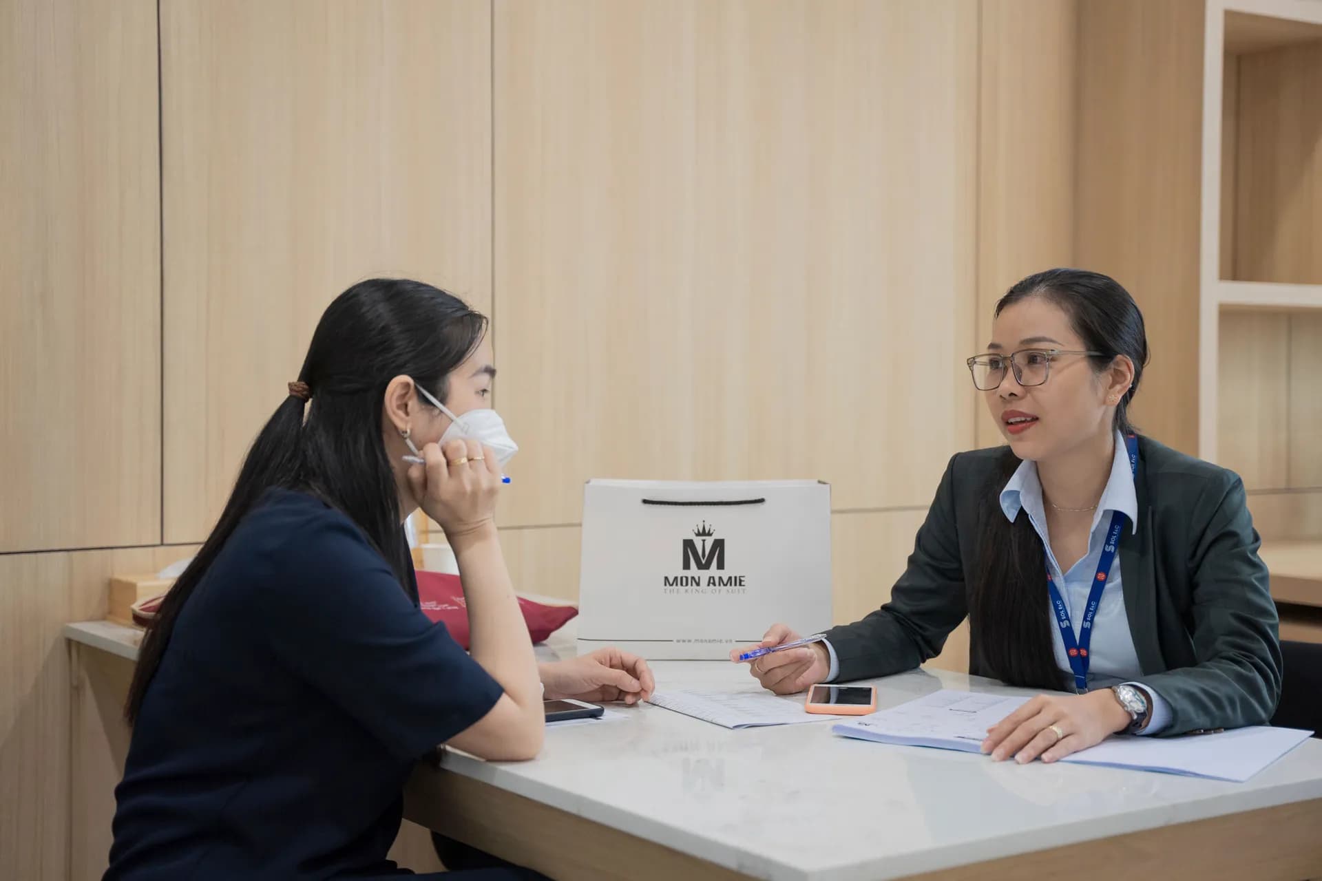 Nhân viên tư vấn Mon Amie trao đổi cùng khách bên bàn trắng, túi giấy có logo