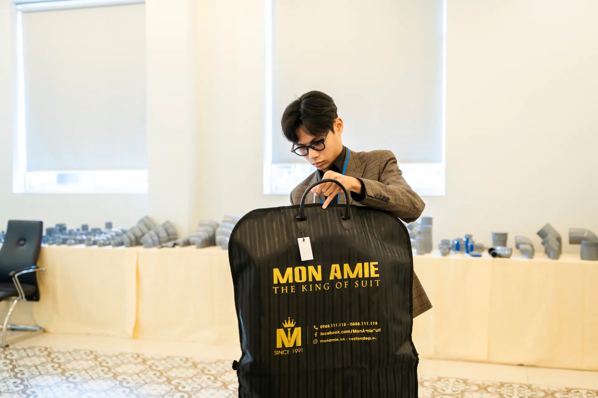 Nhân viên mở túi đựng vest đen chữ vàng Mon Amie, sảnh sáng nền gạch hoa