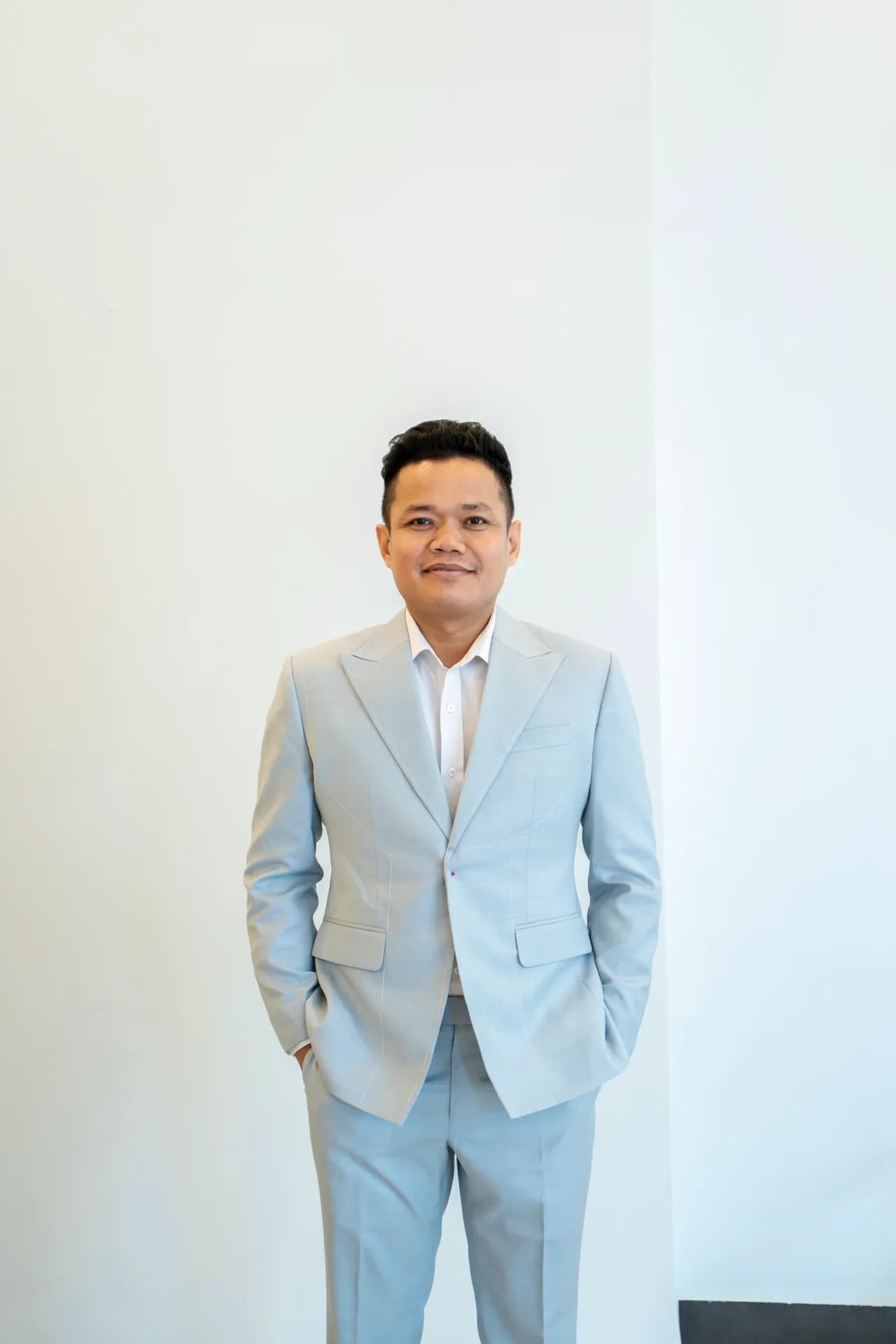 Suit xanh xám nhạt hai khuy ve nhọn, tay đút túi, dáng đứng tự tin trước tường trắng