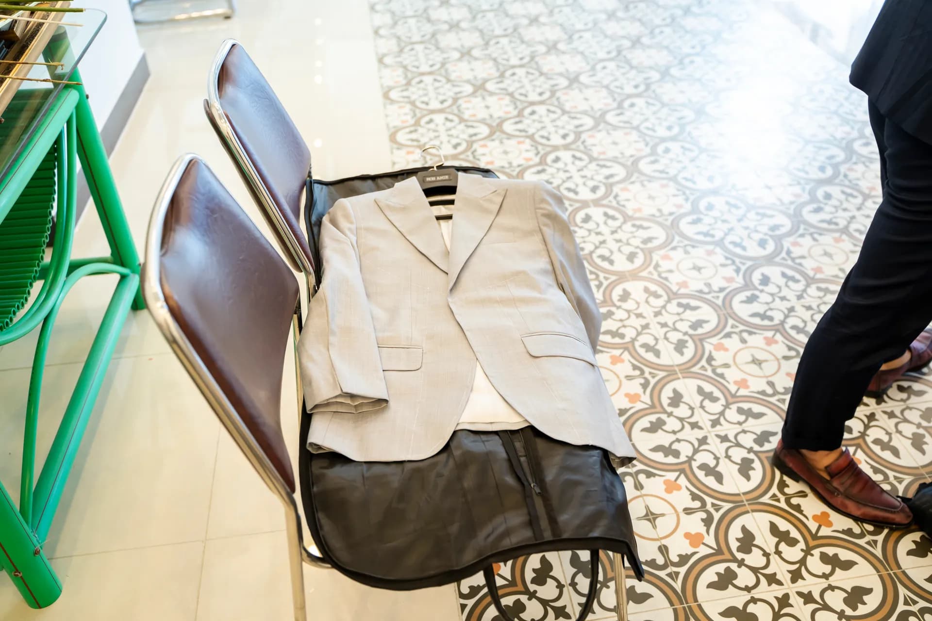 Áo blazer xám nhạt kẻ sọc mảnh đặt trên túi đựng vest, ghế xanh và nền gạch hoa