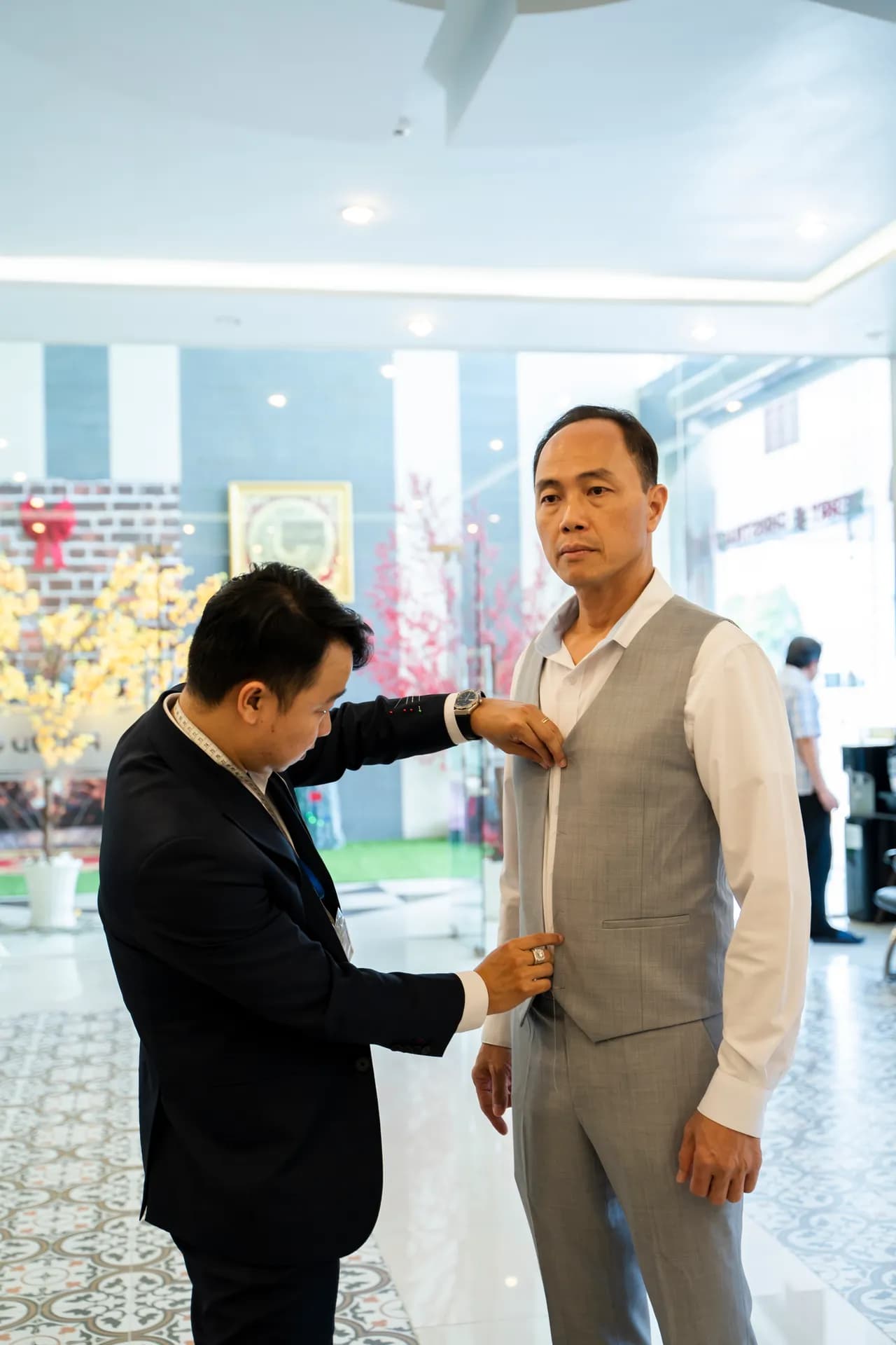 Thợ chỉnh gilê xám caro mảnh cho khách nam trong sảnh sáng, ảnh dọc
