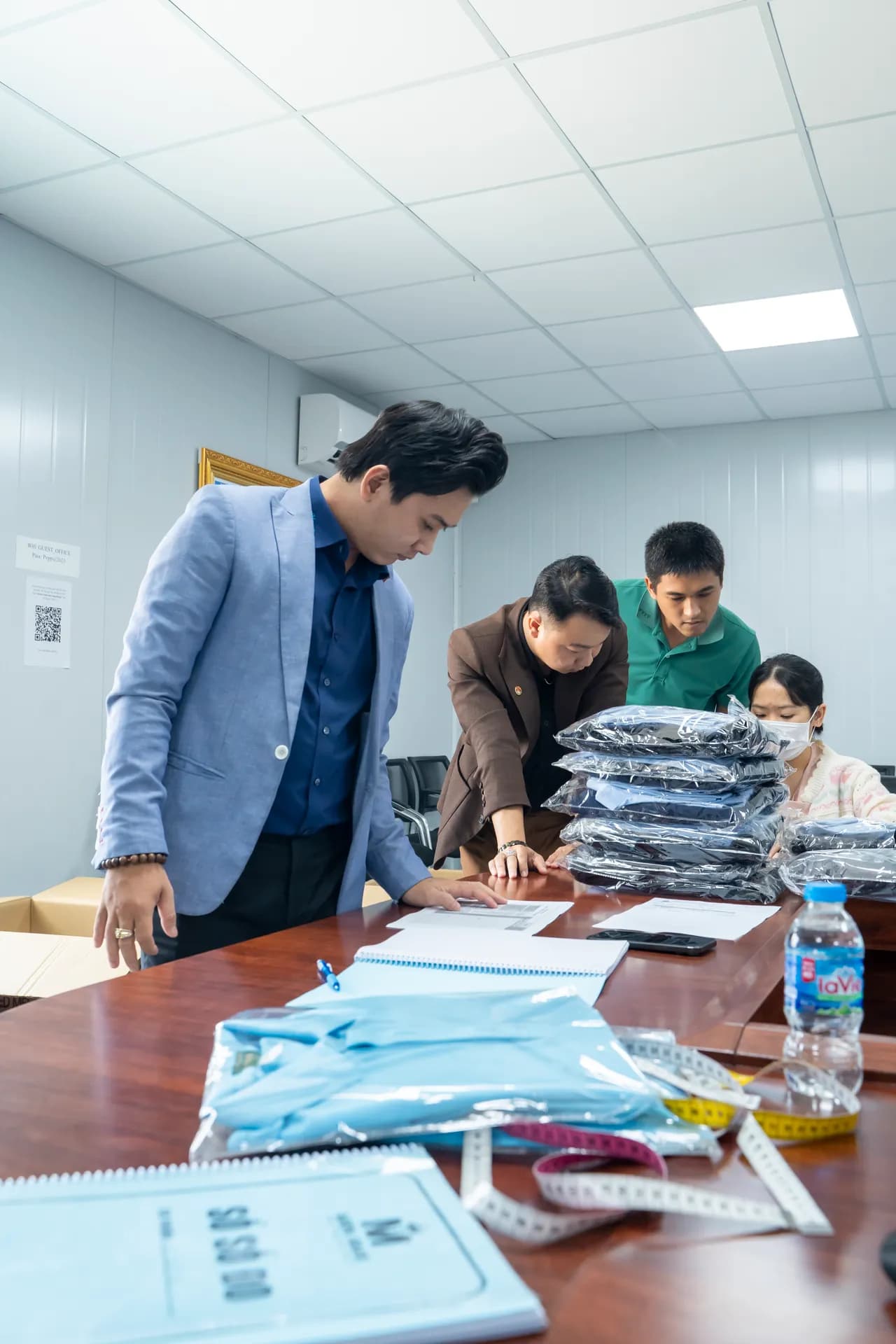 Kiểm đếm áo sơ mi đồng phục xanh nhạt xếp trên bàn họp, thước dây và sổ số đo bên cạnh