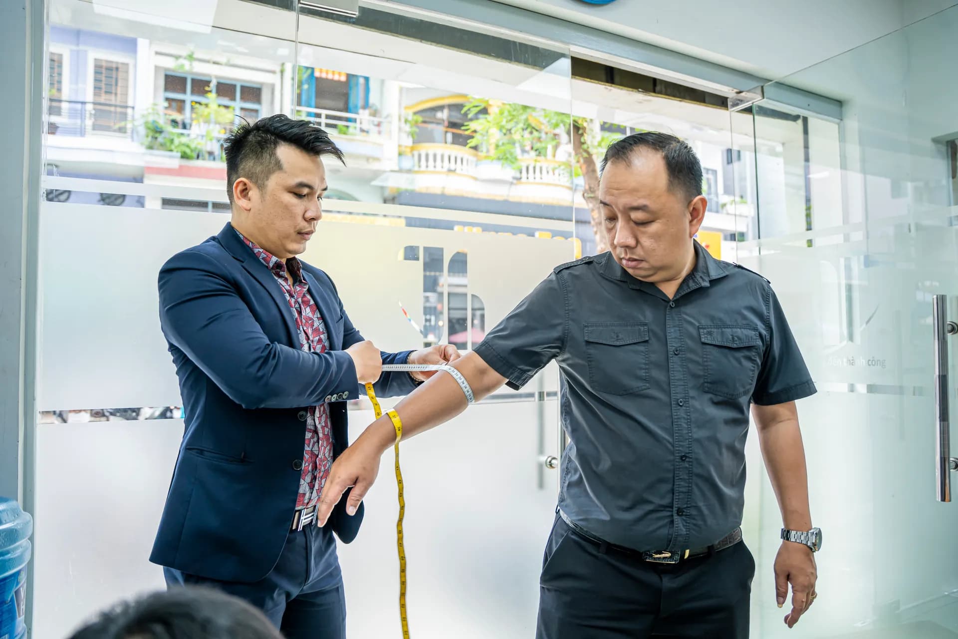Đo vòng bắp tay cho khách áo xám bên cửa kính sáng, đo đồng phục doanh nghiệp