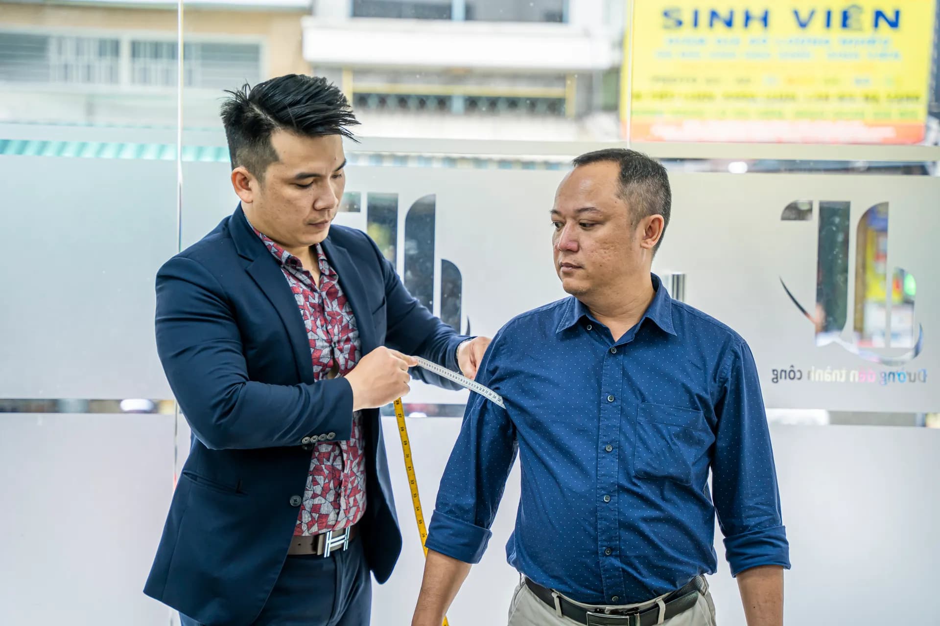 Đo tay áo cho khách sơ mi xanh đậm, thợ mặc blazer navy trao đổi số đo
