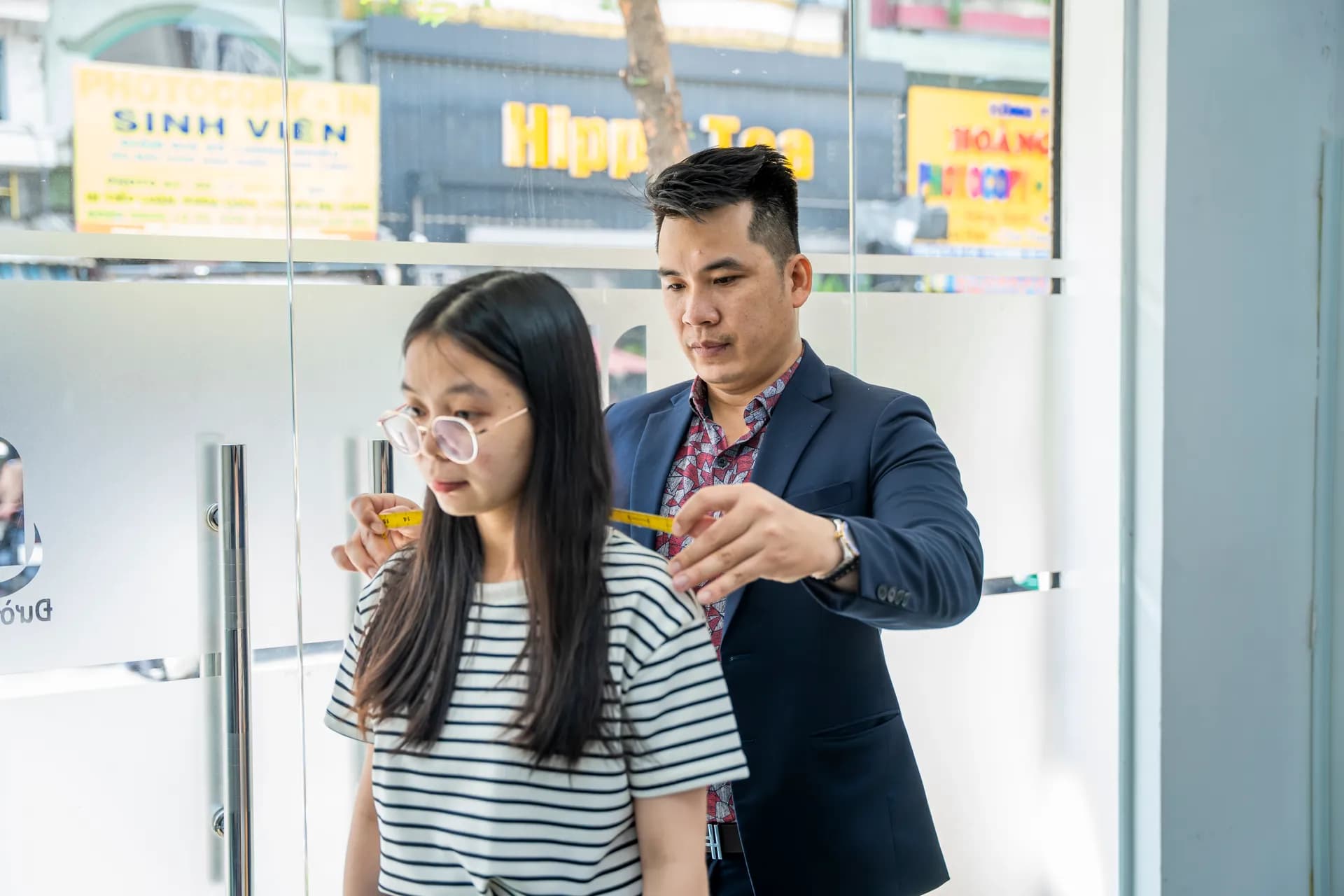 Đo vai cho khách nữ áo sọc ngang, thợ mặc blazer navy, ánh sáng ngoài cửa kính