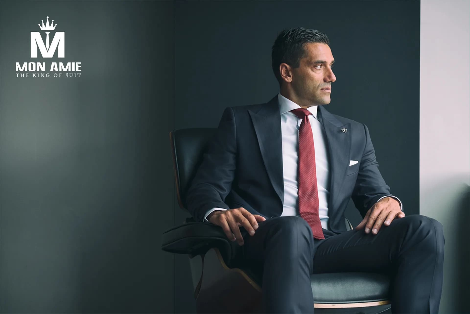 Suit xanh navy, cà vạt đỏ, ngồi ghế da, nền tường xám
