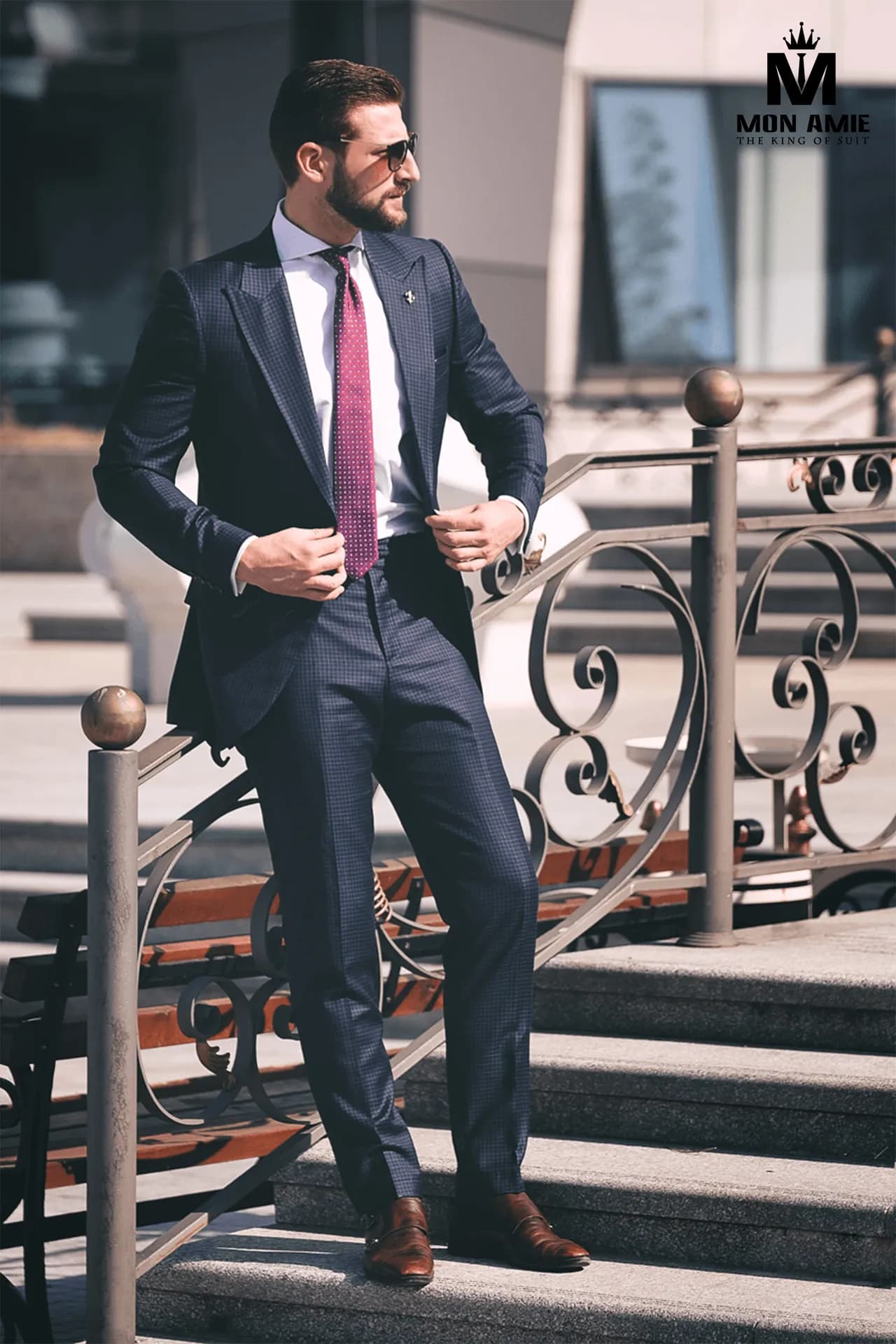 Suit navy cà vạt hồng đỏ, một tay chỉnh khuy áo, đứng bên lan can sắt uốn