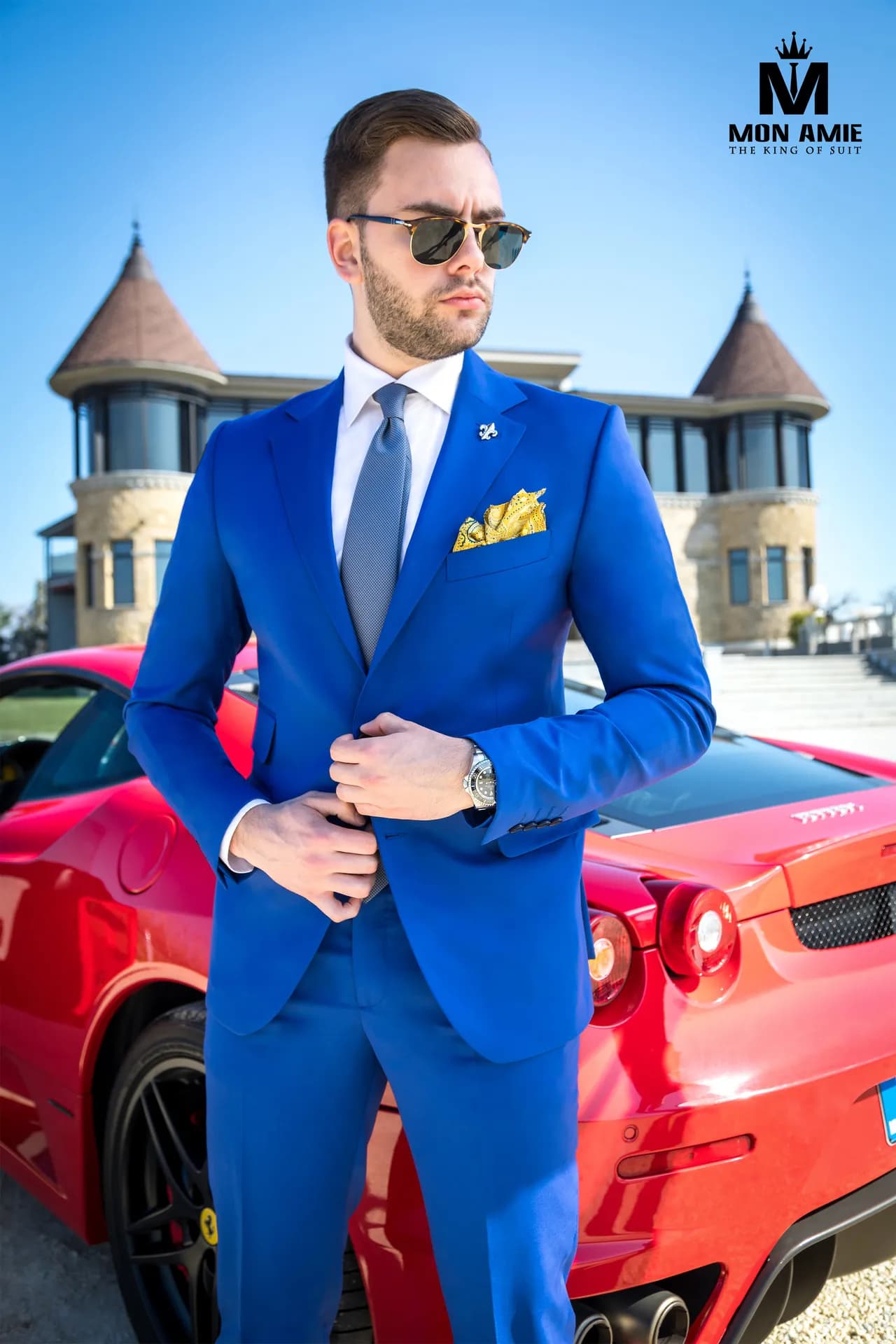 Suit xanh cobalt, kính râm, khăn cài túi vàng, đứng cạnh xe thể thao đỏ