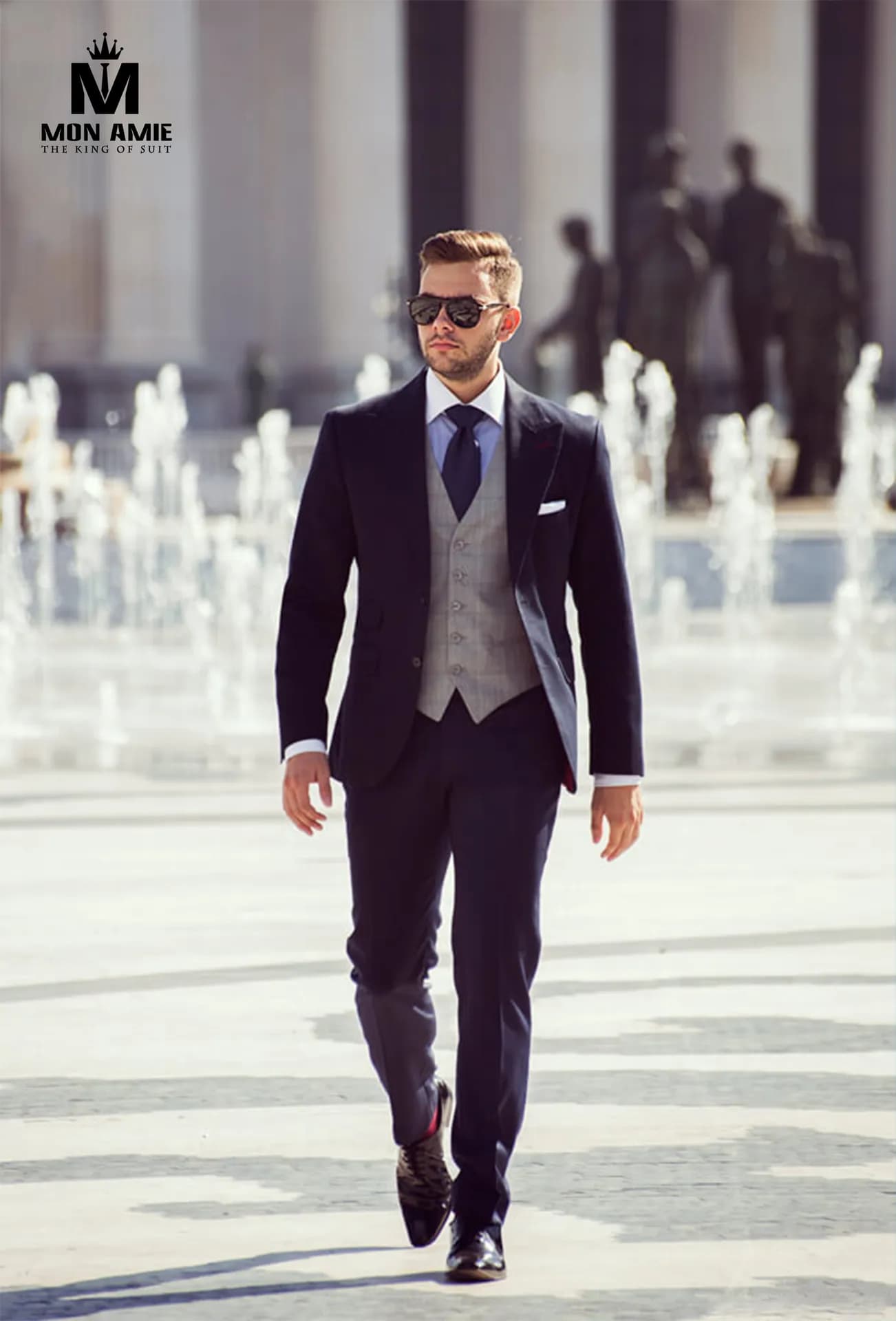 Suit navy gilê xám, kính râm, bước đi giữa quảng trường đài phun nước
