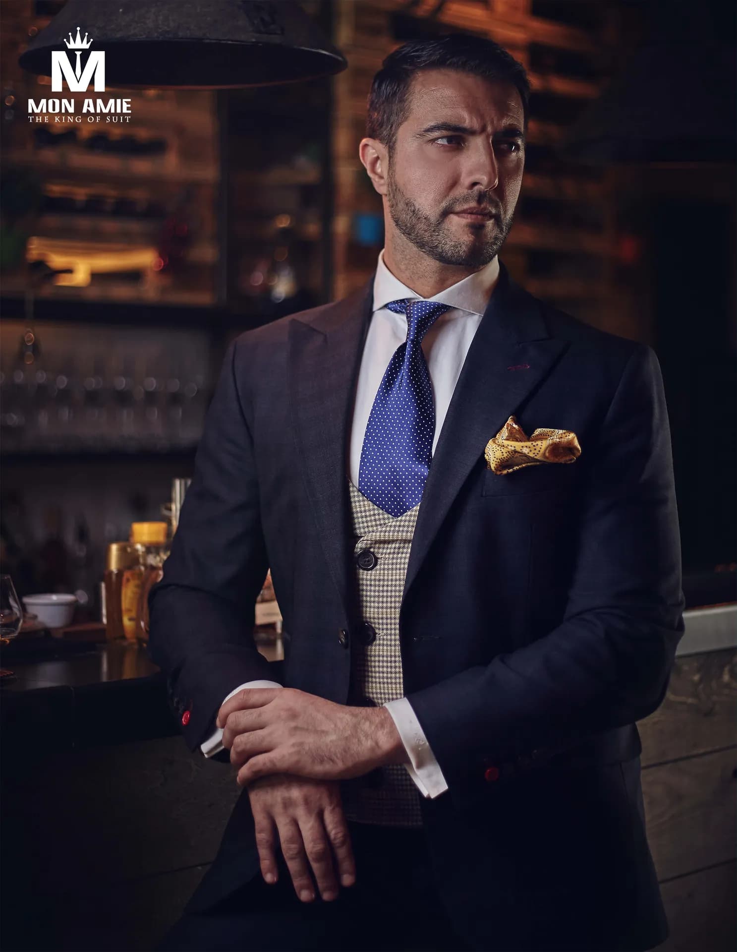 Suit xanh navy gilê kẻ nhỏ, cà vạt xanh, khăn cài túi vàng, nền quầy bar tối