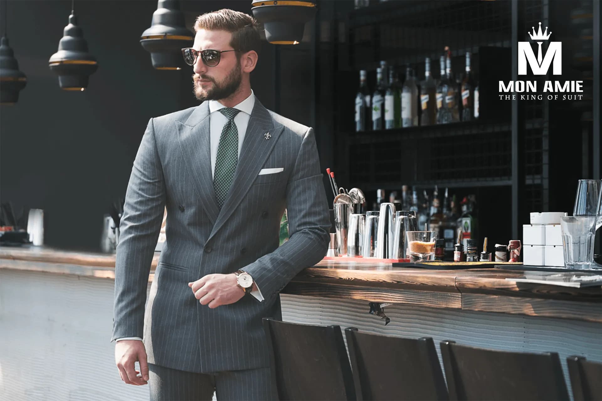 Suit xám kẻ sọc hai hàng khuy, cà vạt xanh lá, kính râm bên quầy bar