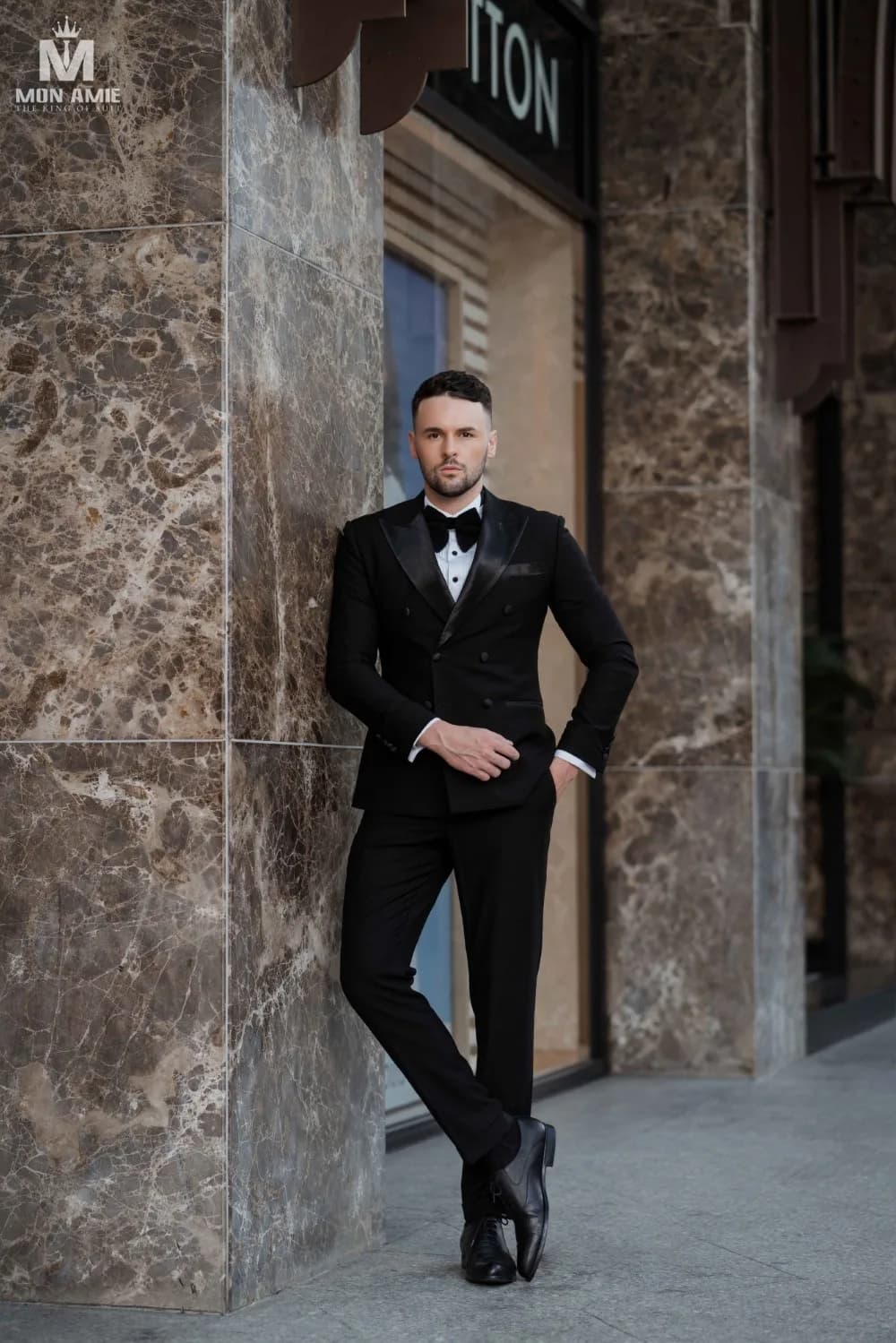 Tuxedo đen tựa cột đá cẩm thạch, giày da bóng và nơ đen
