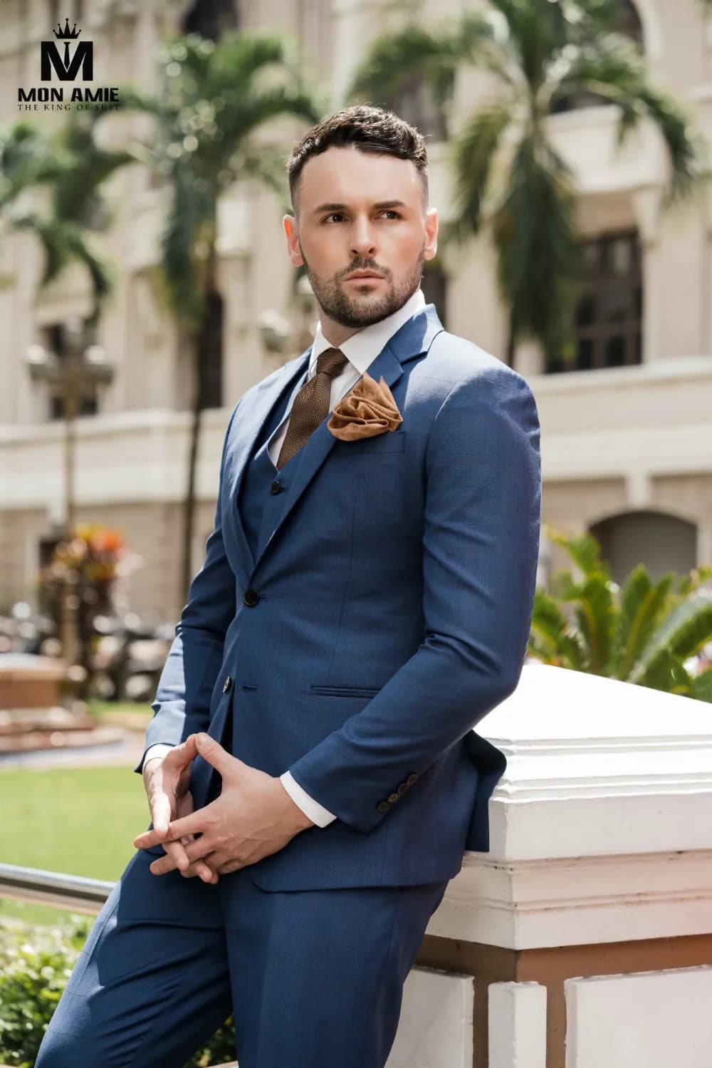 Suit xanh navy ba mảnh, đứng dựa lan can giữa hàng cọ