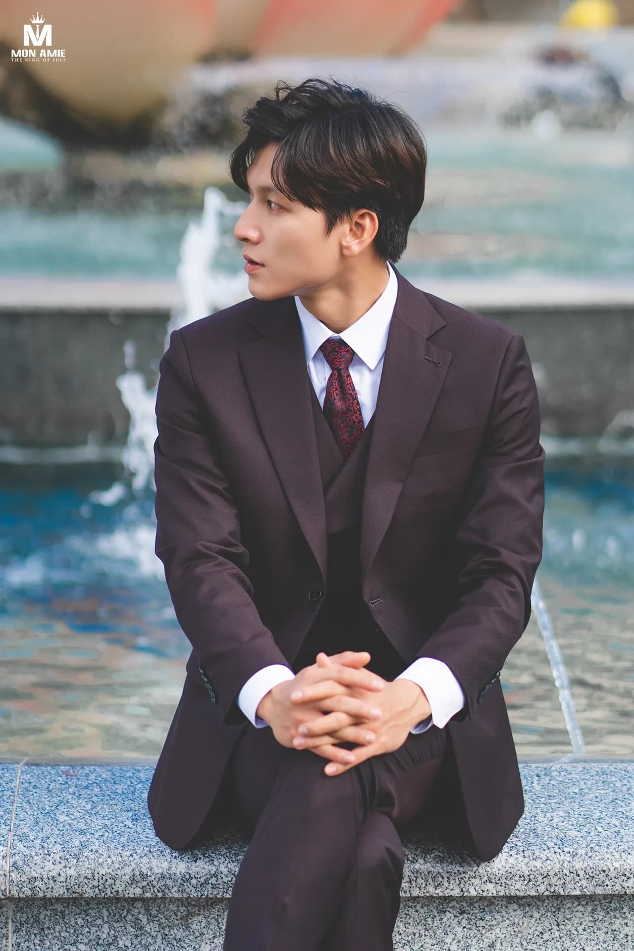 Suit ba mảnh nâu tím than, cà vạt đỏ hoa, ngồi nhìn nghiêng bên đài phun nước