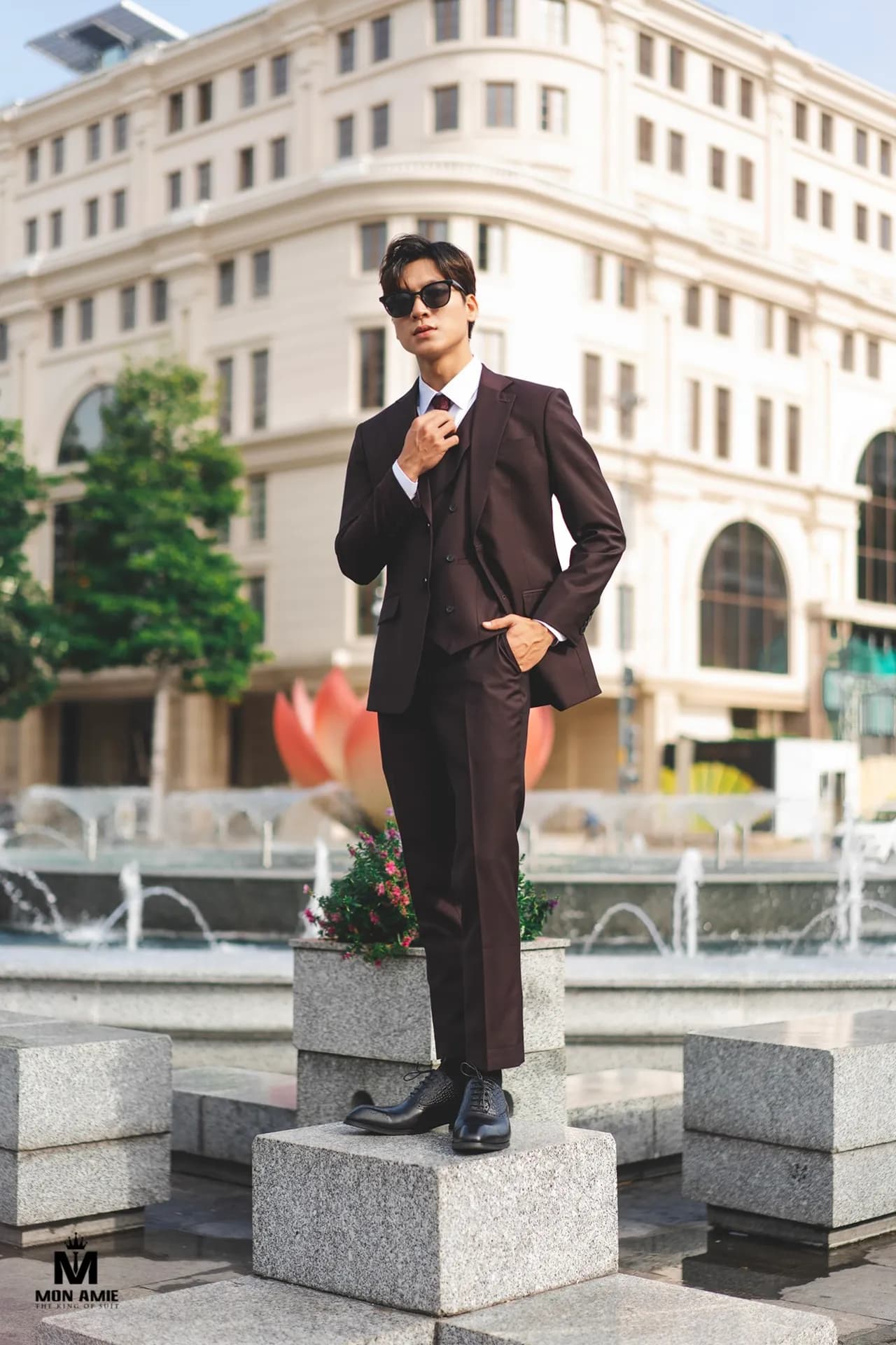 Suit ba mảnh nâu tím than, kính râm, đứng trên bệ đá trước toà nhà cổ
