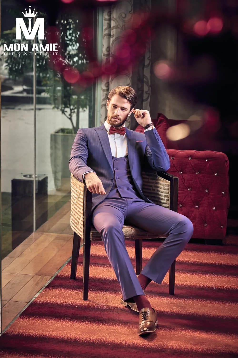 Suit ba mảnh xanh sọc mảnh, gilet cài hai hàng, nơ đỏ mận, ngồi thư thái giữa sắc tím