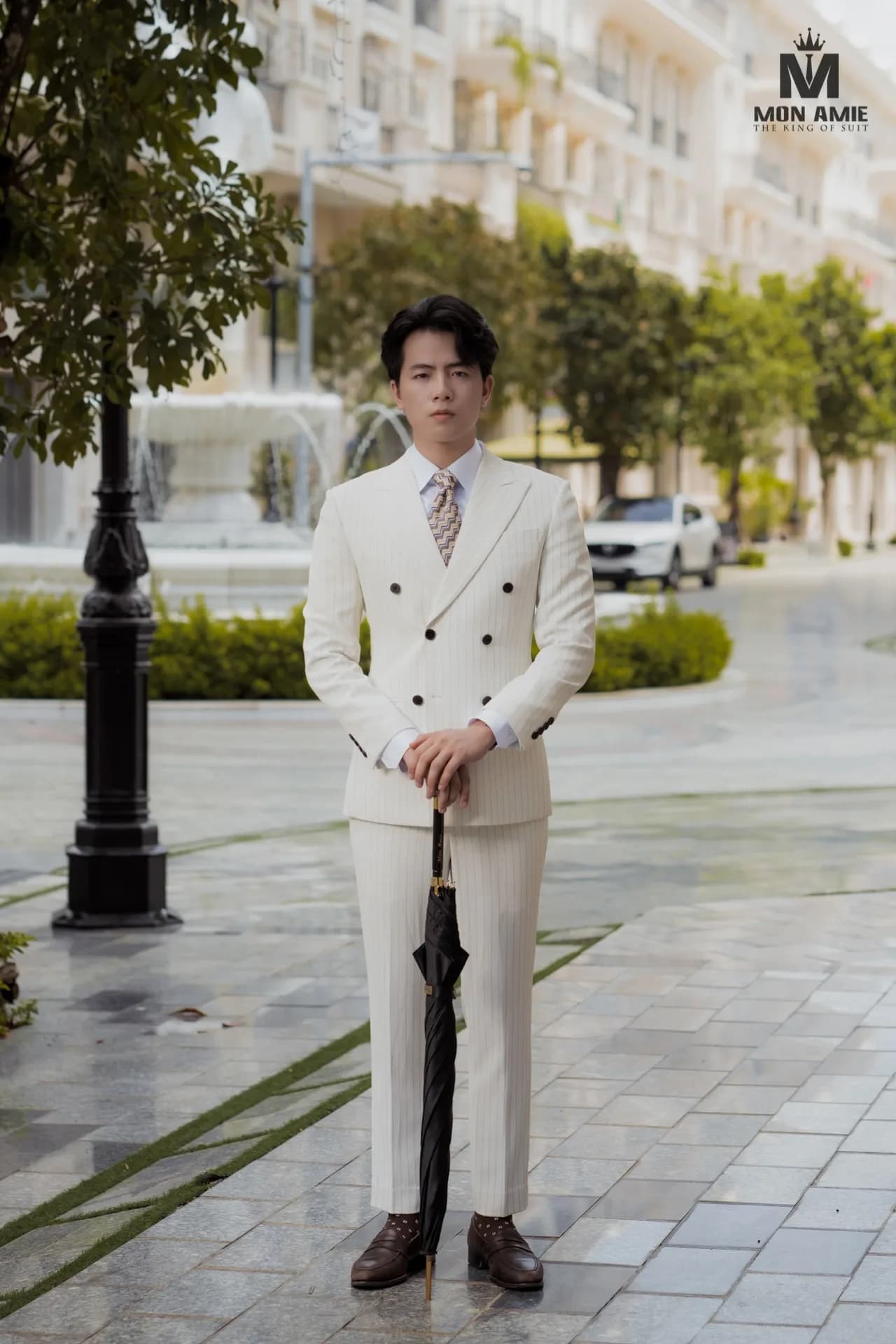Suit trắng kem kẻ sọc hai hàng cúc, cầm ô đen giữa phố lát đá và đài phun nước