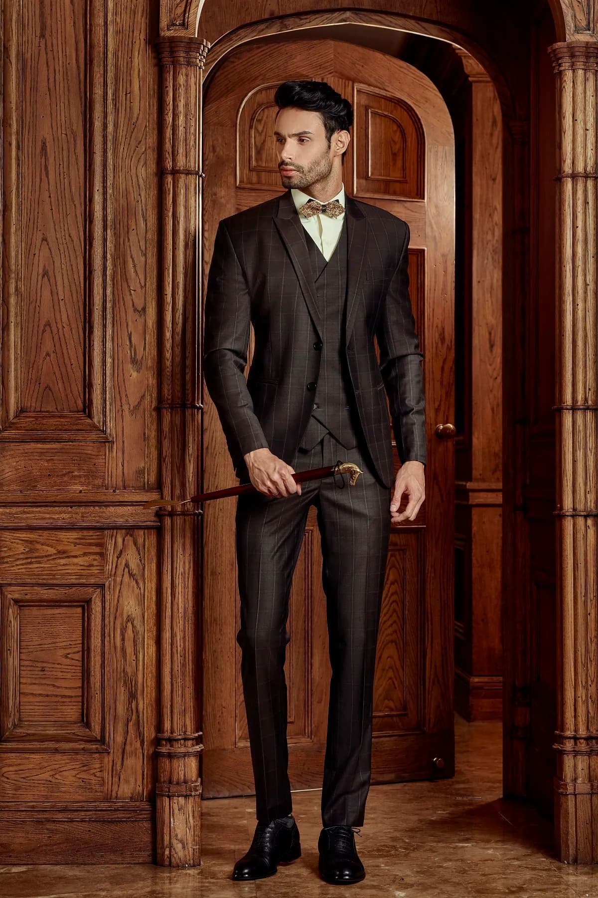 Suit ba mảnh nâu đậm, nơ vàng, cầm ba-toong, đứng trước cửa vòm gỗ