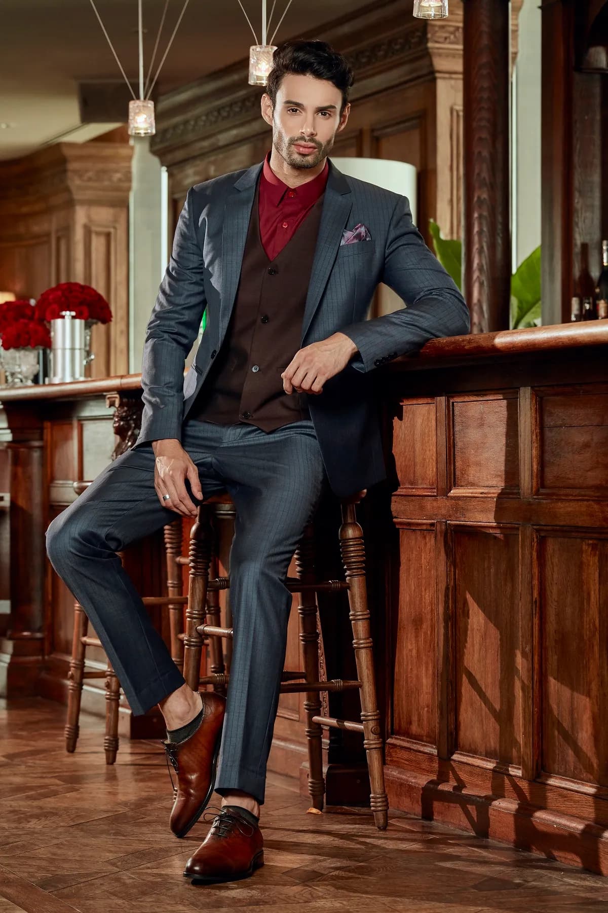 Suit xám xanh phối gile nâu, sơ mi đỏ đô, ngồi ghế cao tại quầy bar gỗ