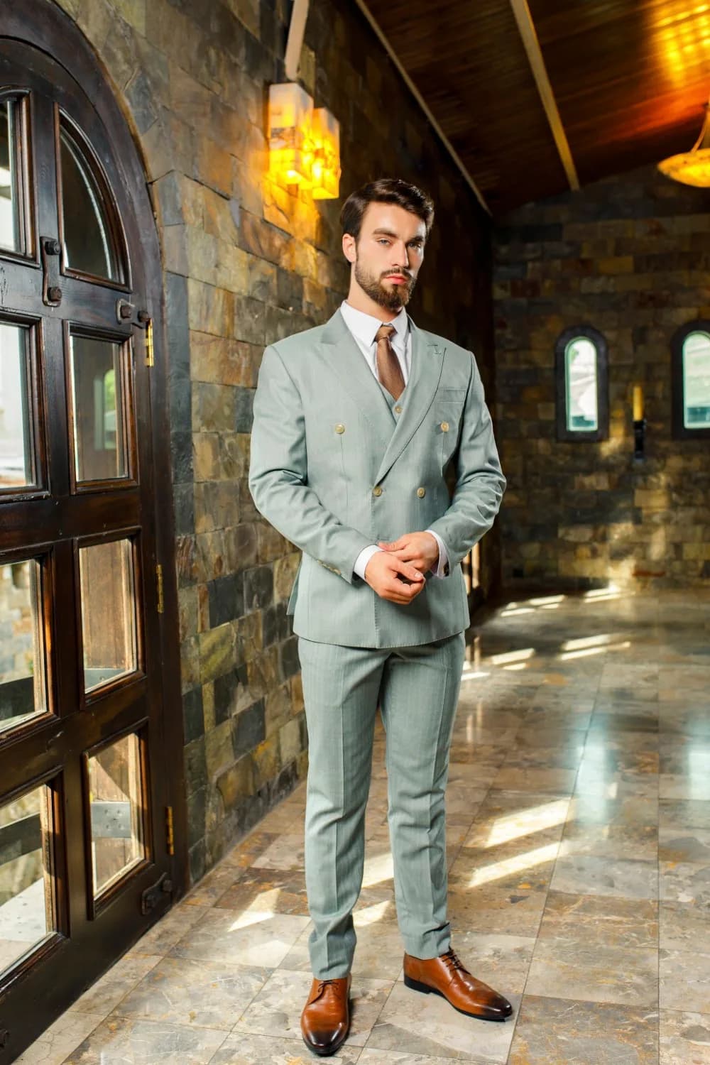 Bộ suit ba mảnh xanh sage cài hai hàng, giày da nâu, sảnh đá cổ kính ánh nắng