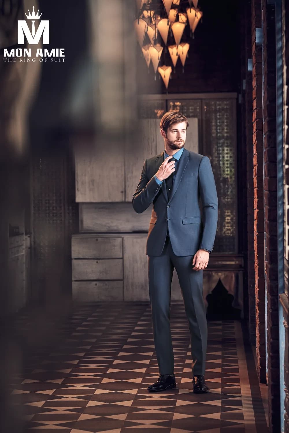 Suit xanh xám ba mảnh, tay chỉnh cà vạt, dáng đứng thanh lịch trên nền gạch hoa cổ điển