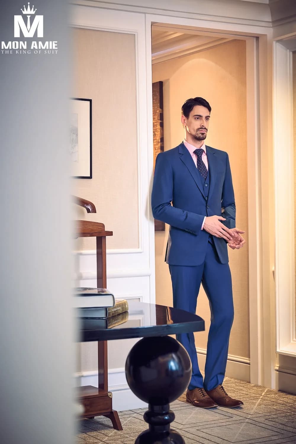 Suit xanh cobalt ba mảnh, sơ mi hồng nhạt, cà vạt chấm bi, đứng nơi cửa ánh vàng