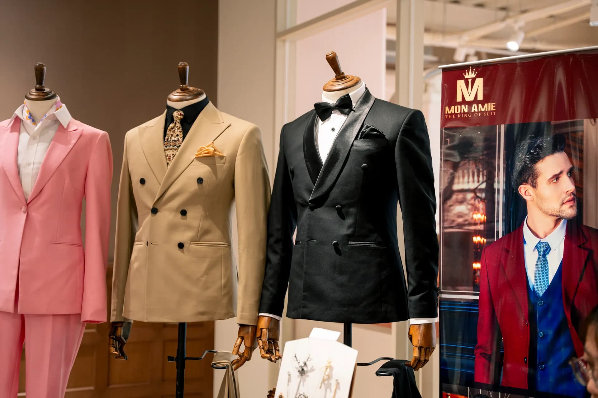 Ba manơcanh vest hồng, be cài hai hàng khuy và tuxedo đen cạnh banner Mon Amie