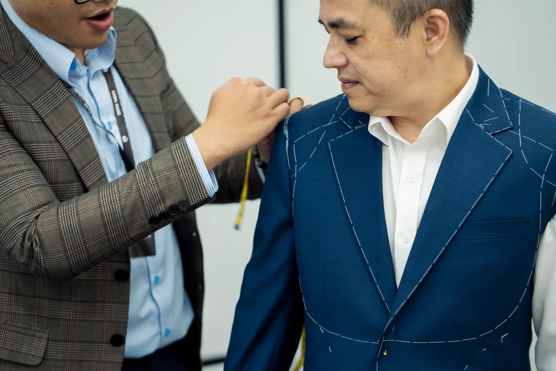 Thợ may đo vai cho khách đang thử vest xanh lược chỉ, thước dây vàng