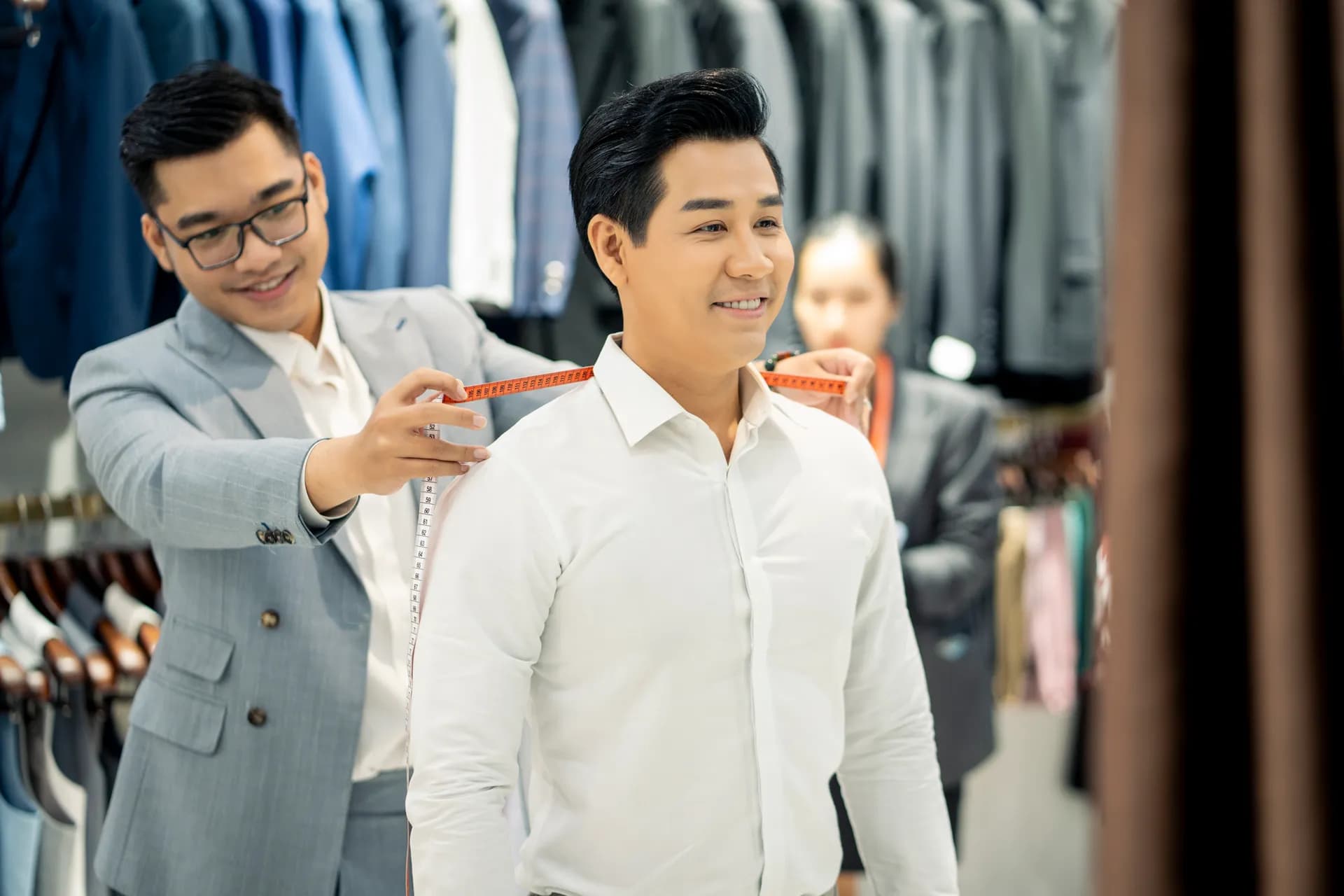Thợ may đo vai cho khách sơ mi trắng đang mỉm cười giữa giá vest trong showroom