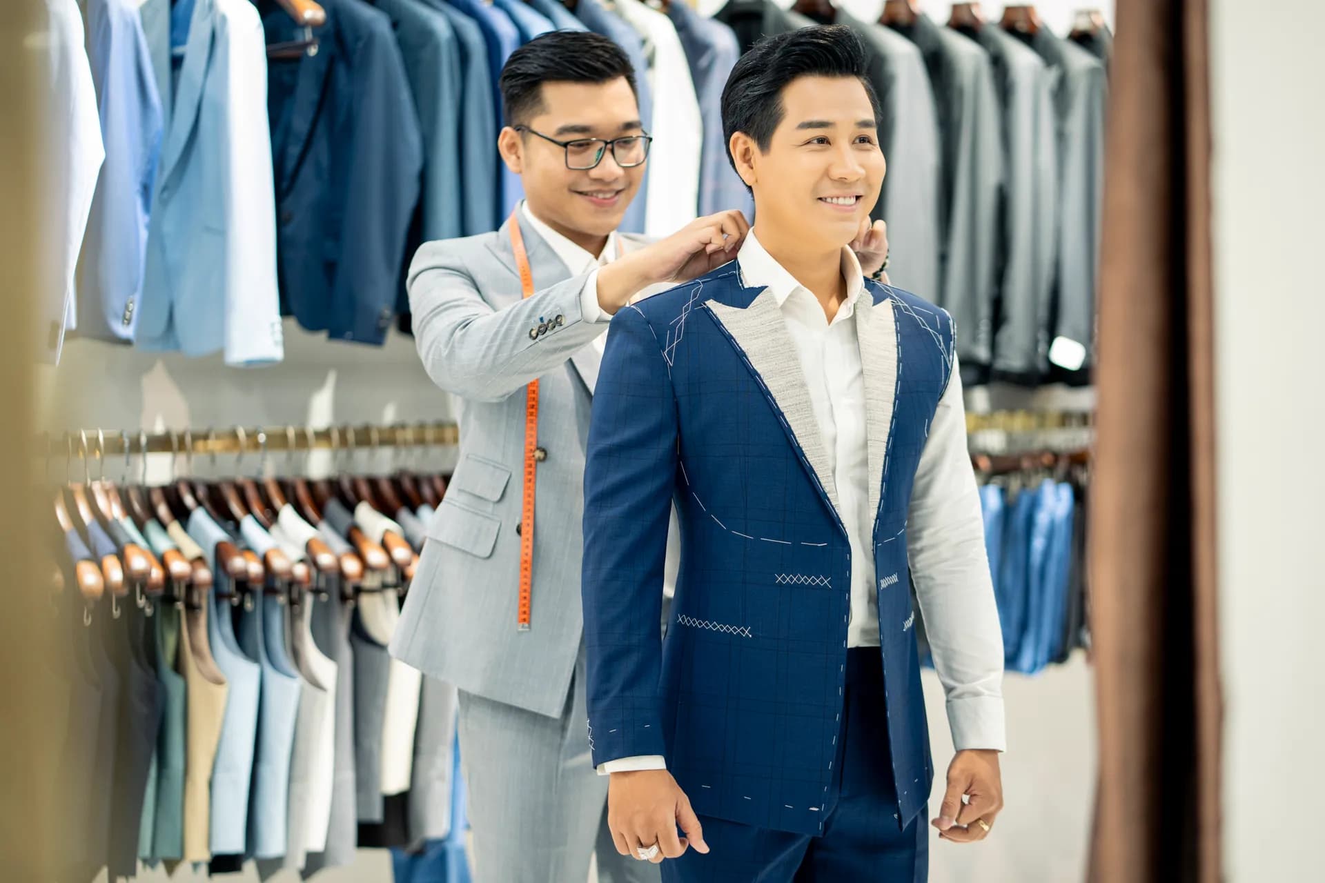 Thợ may chỉnh cổ vest xanh còn lược chỉ trên người khách đang cười tươi