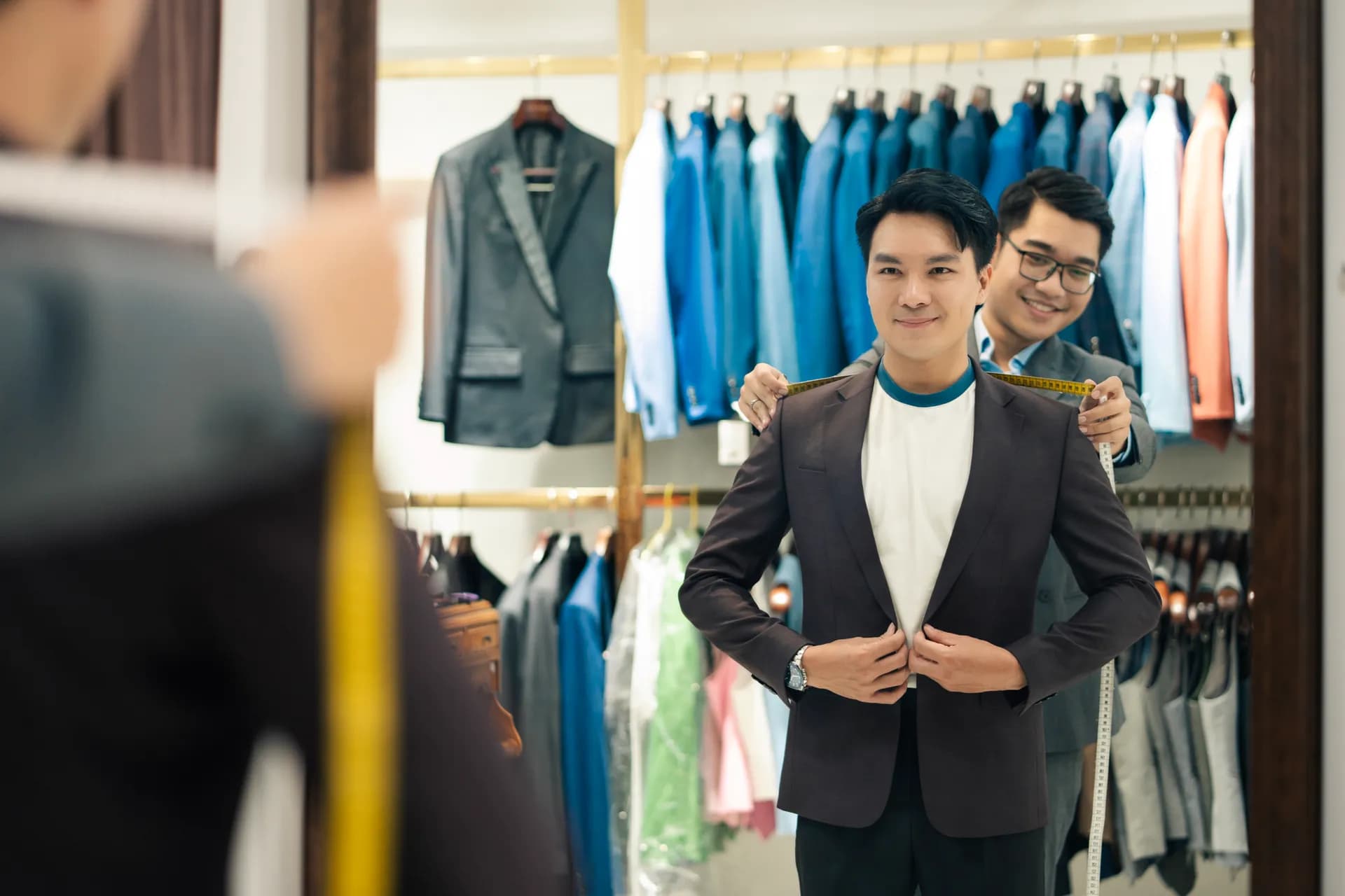 Khách cười tươi thử áo vest nâu tối, thợ may đo vai qua gương trong showroom