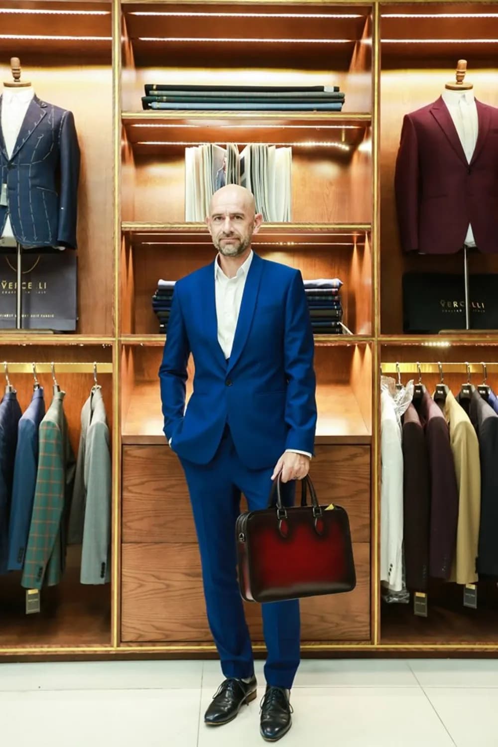 Suit xanh cobalt, tay đút túi, xách cặp da đỏ rượu trước kệ vest