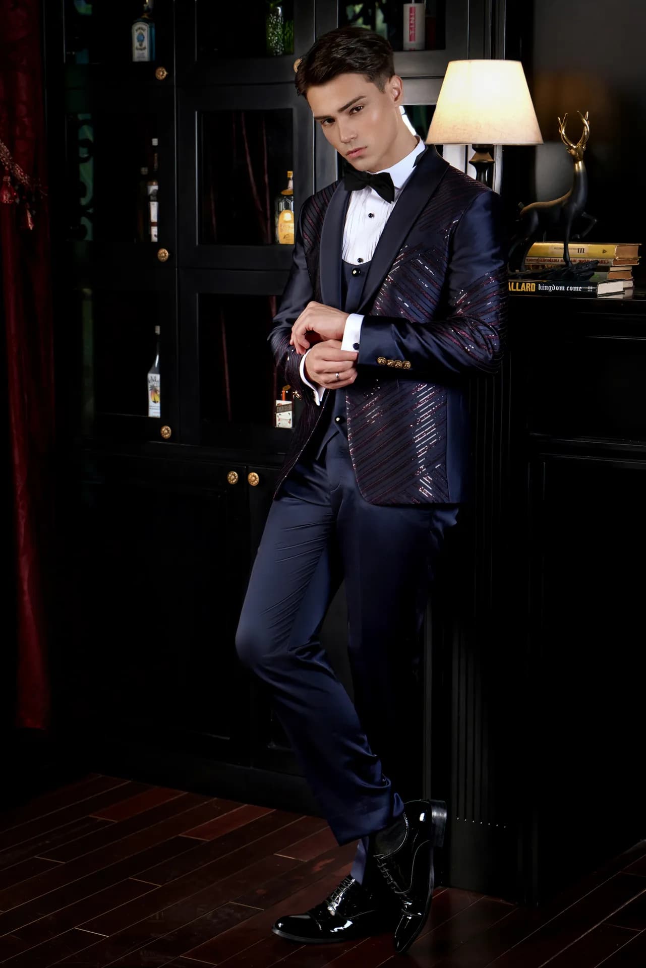 Suit navy dệt kim tuyến ánh đỏ ba mảnh, nơ đen, tựa kệ gỗ cổ điển.