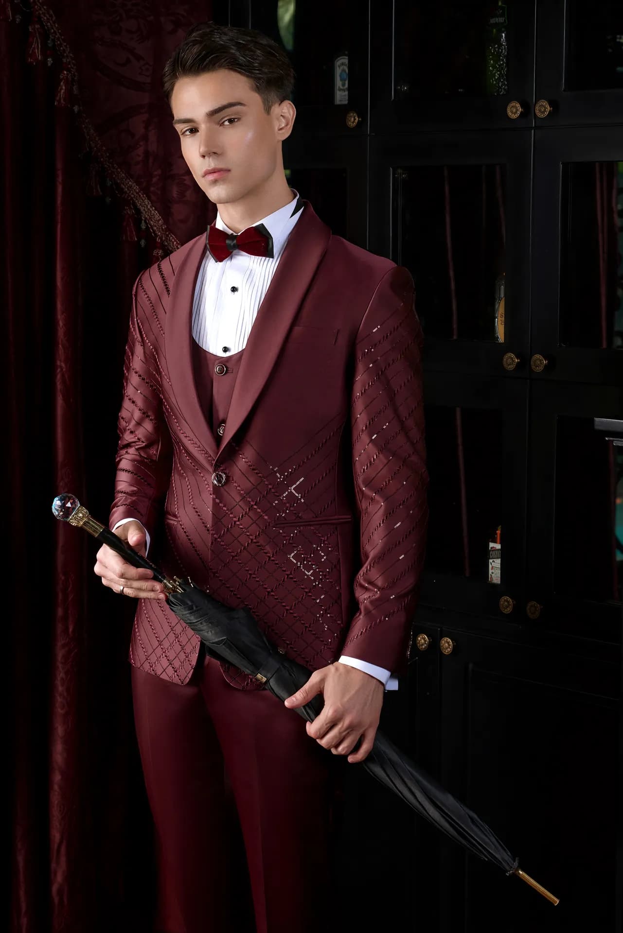 Suit đỏ đô ba mảnh, nơ đen, cầm ô đen trước rèm nhung đỏ