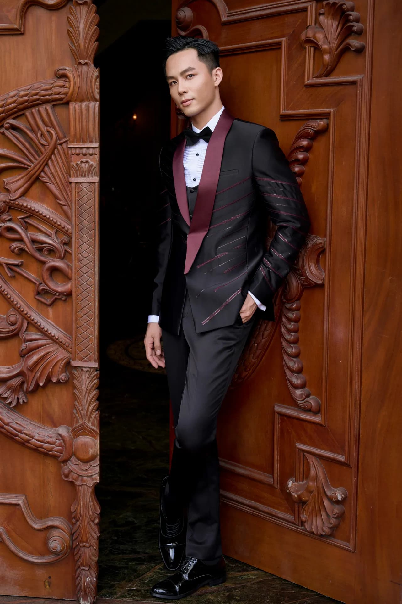 Tuxedo đen cổ shawl đỏ đô, tựa cánh cửa gỗ chạm khắc