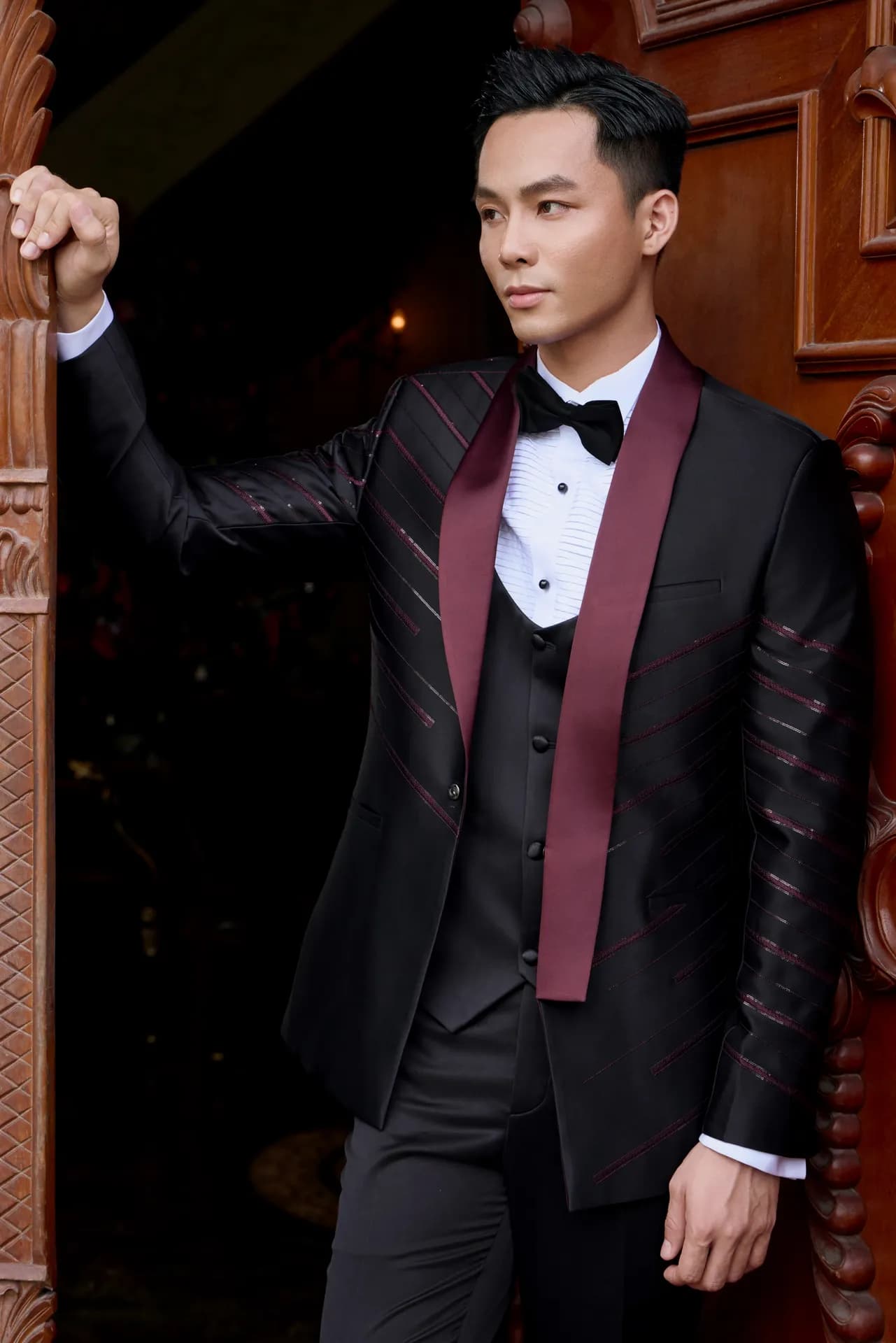 Tuxedo đen cổ shawl đỏ đô, tay chống cửa gỗ chạm khắc